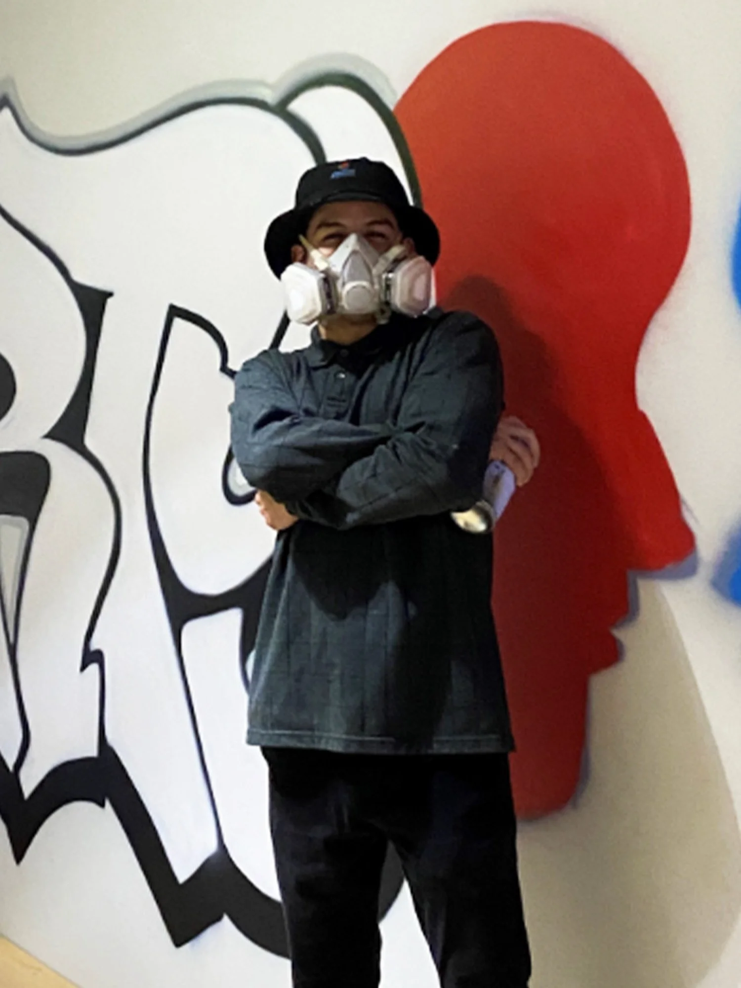 A man with a hat and respirator mask holding a spray paint can and standing in front of a mural.