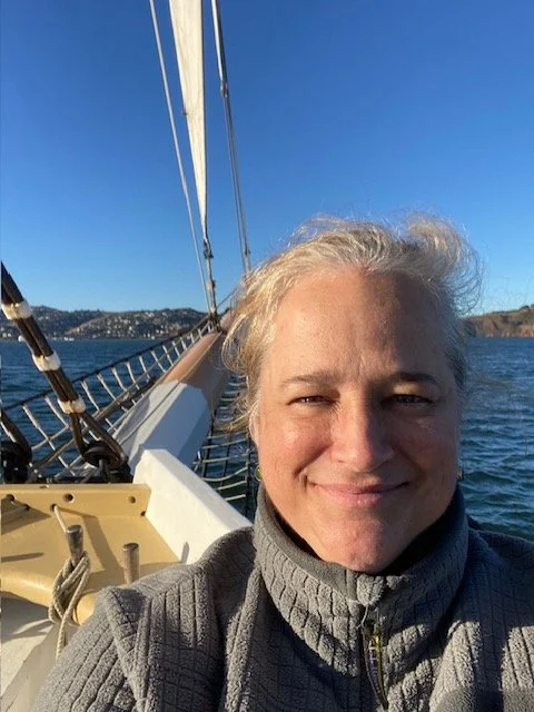 Person taking a selfie on a sailboat with water, coastline, and blue sky in the background.