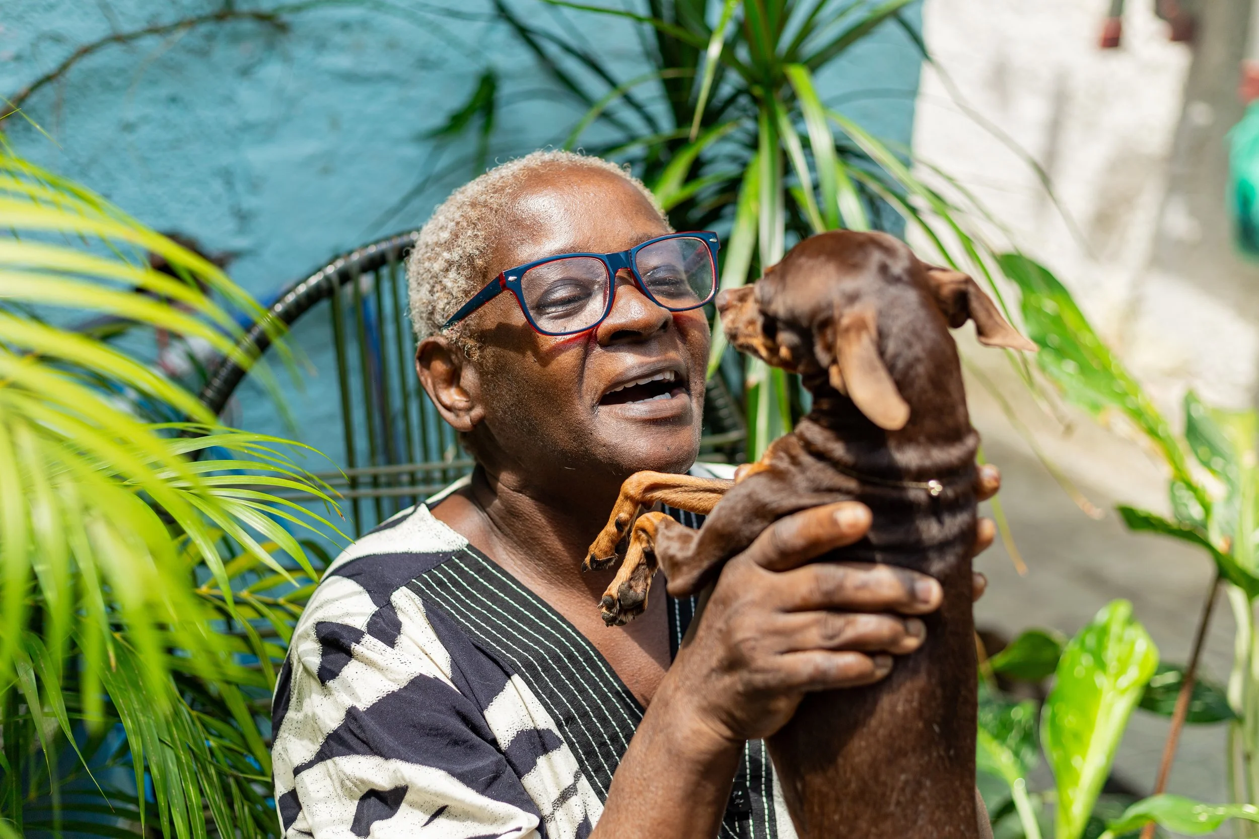 An elderly woman wearing glasses is holding a small brown dog outdoors surrounded by green plants and a blue wall.