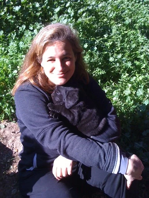 Woman outdoors holding a black puppy with green foliage in the background.