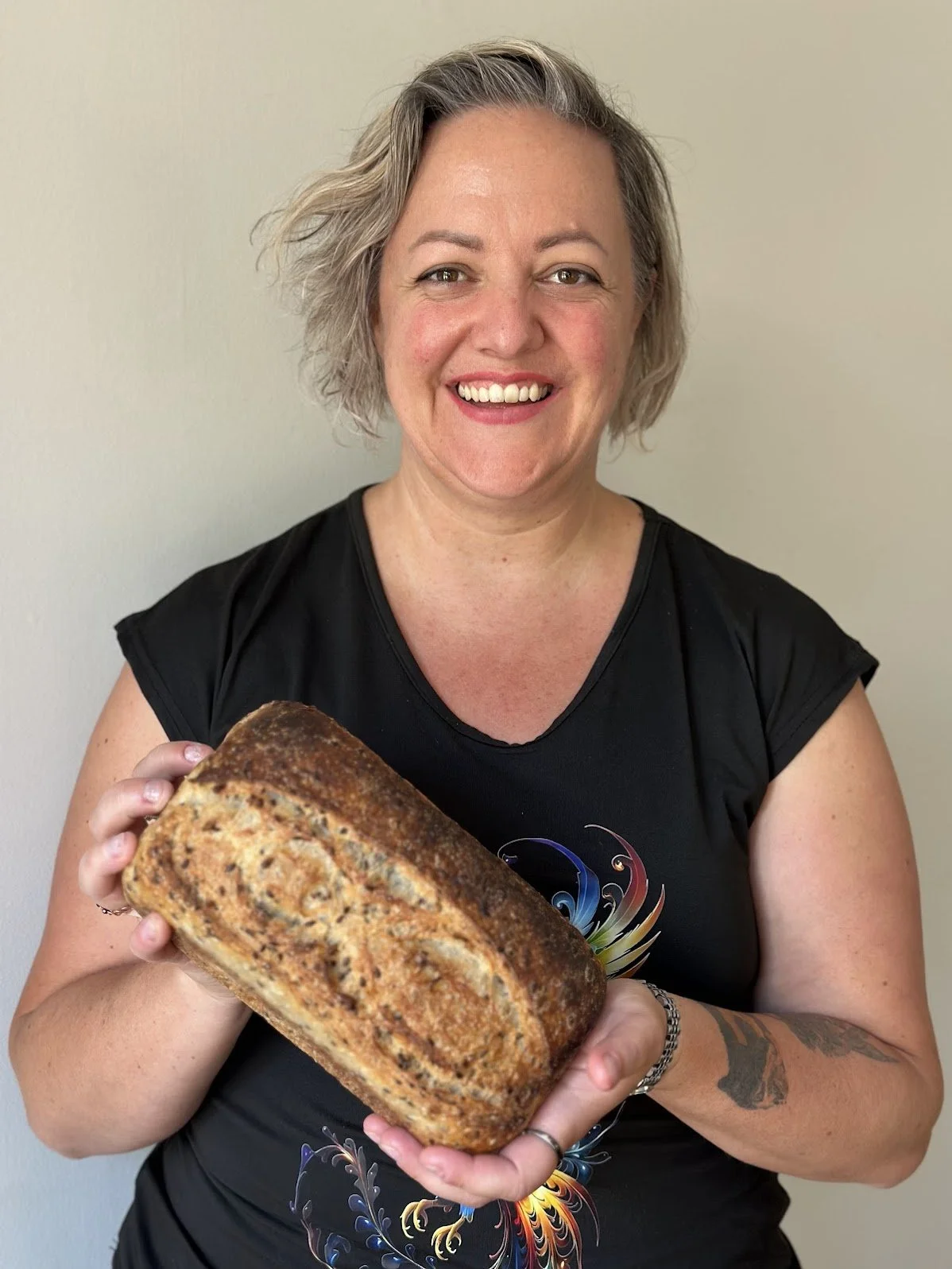 Kapiti Delicious Doughs Owner Meg Price is smiling and holding a freshly baked sunflower and linseed sourdough loaf.