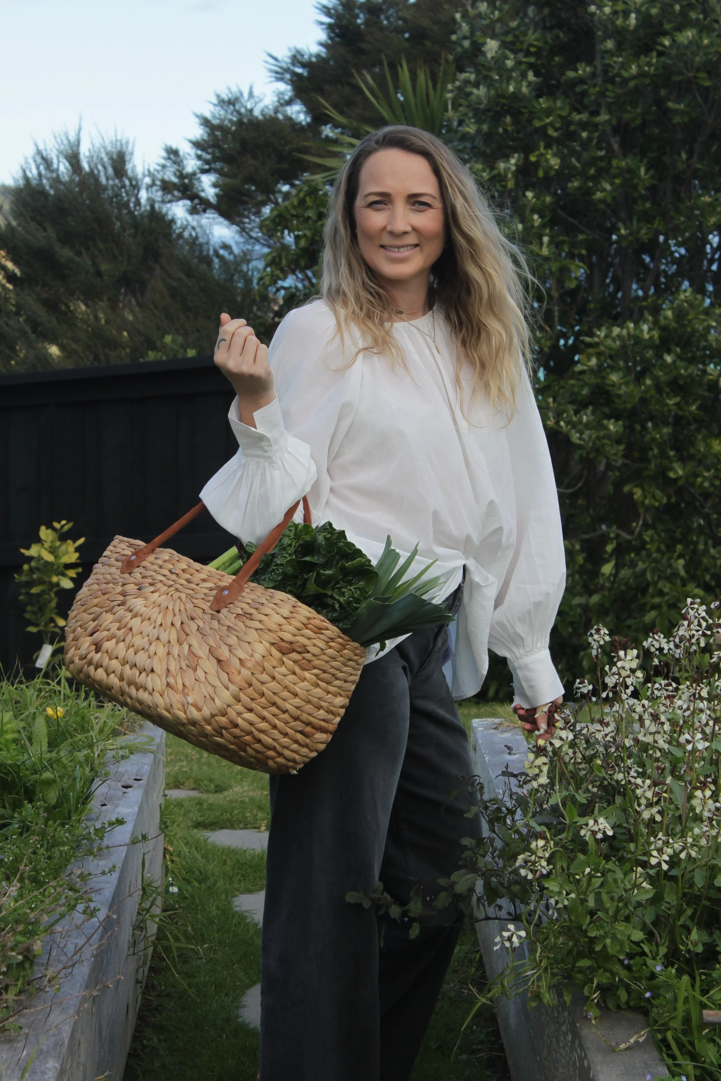 Woman with long hair smiling, holding a woven basket filled with green vegetables, standing in a garden surrounded by plants and flowers.