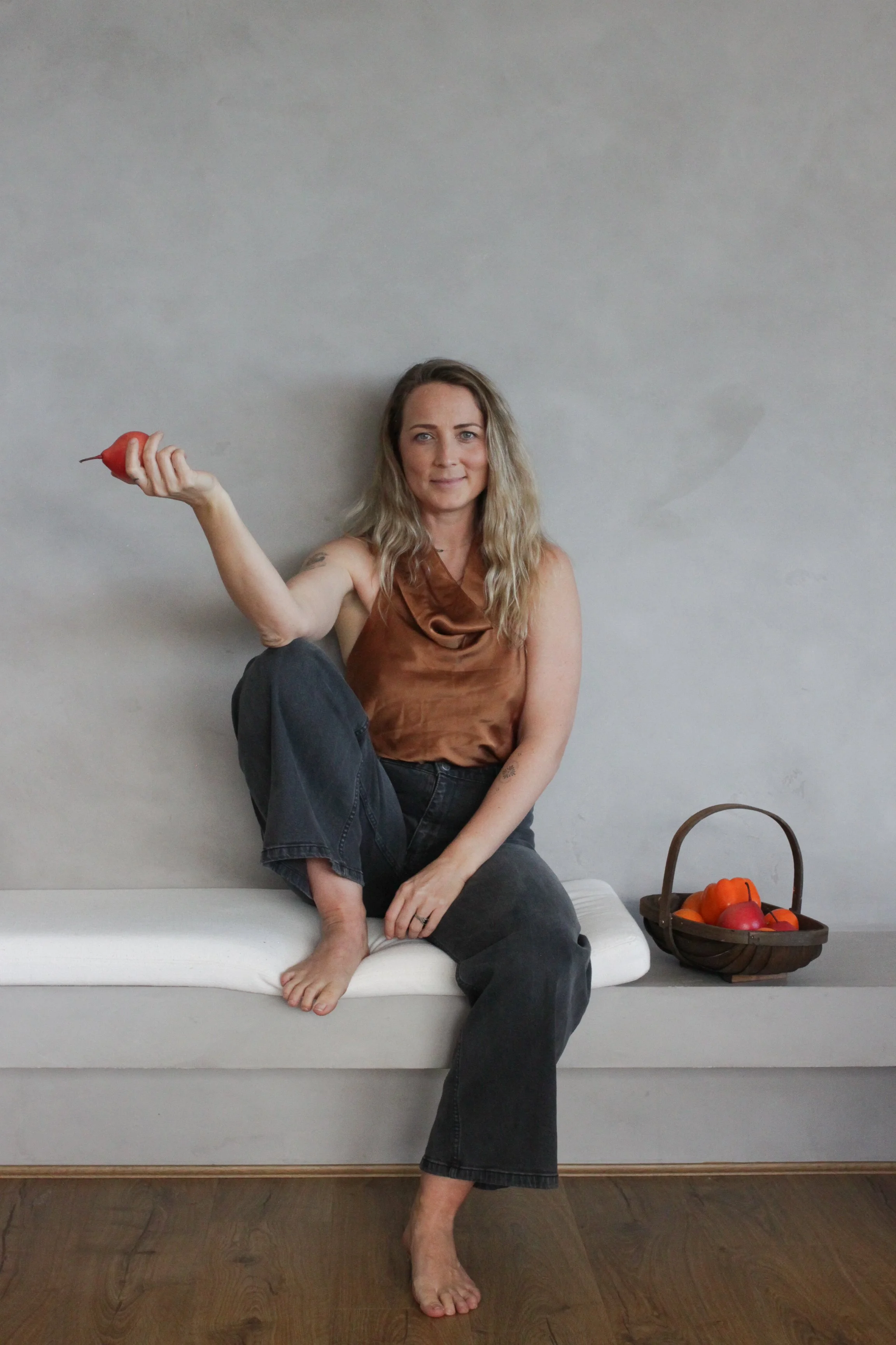 A woman with wavy blonde hair sitting on a white padded surface in front of a plain gray wall, holding a red object in her right hand with her arm extended. She is wearing a brown sleeveless top and dark jeans, with one leg crossed over the other and barefoot. There is a basket of colorful vegetables on the right side of her.