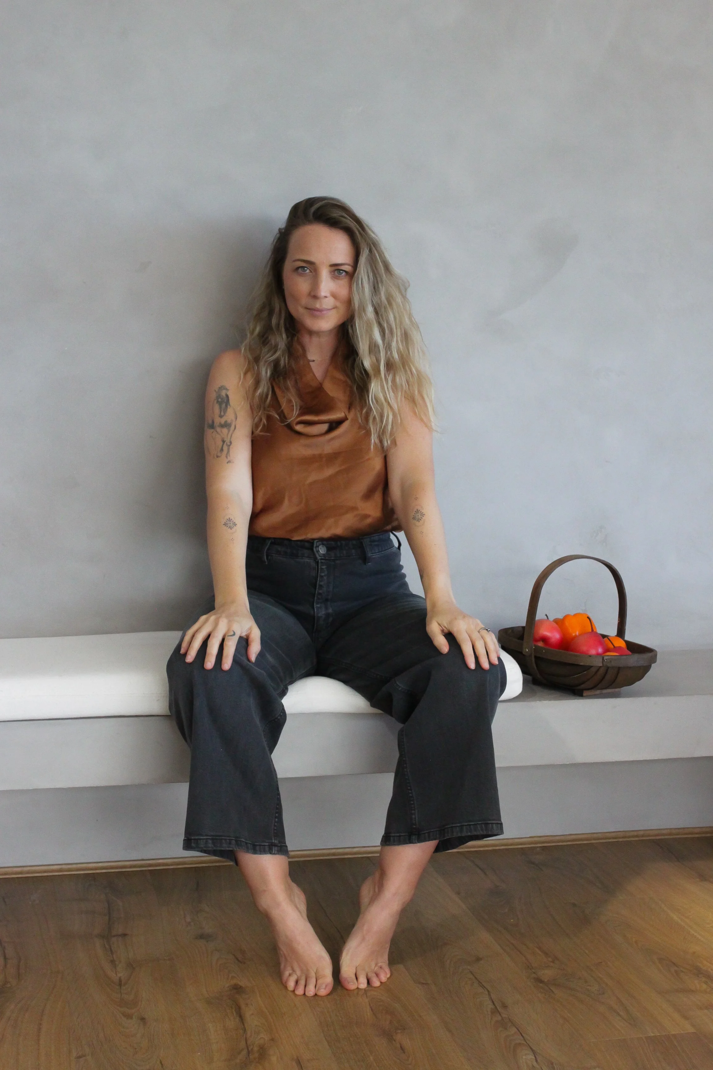 A woman with wavy blonde hair sitting on a white bench, wearing a brown sleeveless top and black jeans, with a basket of apples and orange bell peppers beside her, against a gray background.