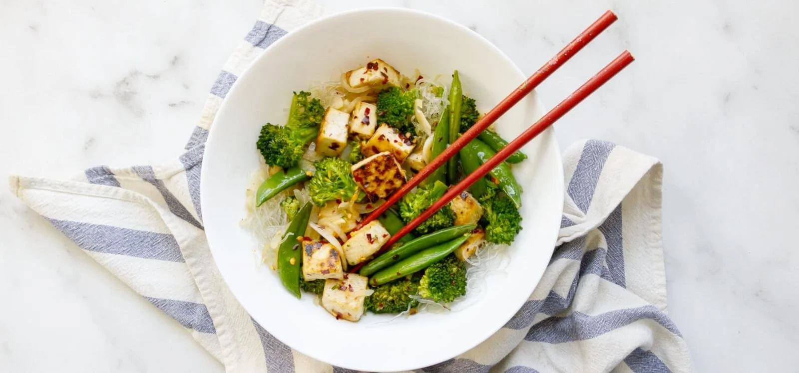 A white bowl of Asian stir-fried vegetables and tofu on a cloth napkin with chopsticks.