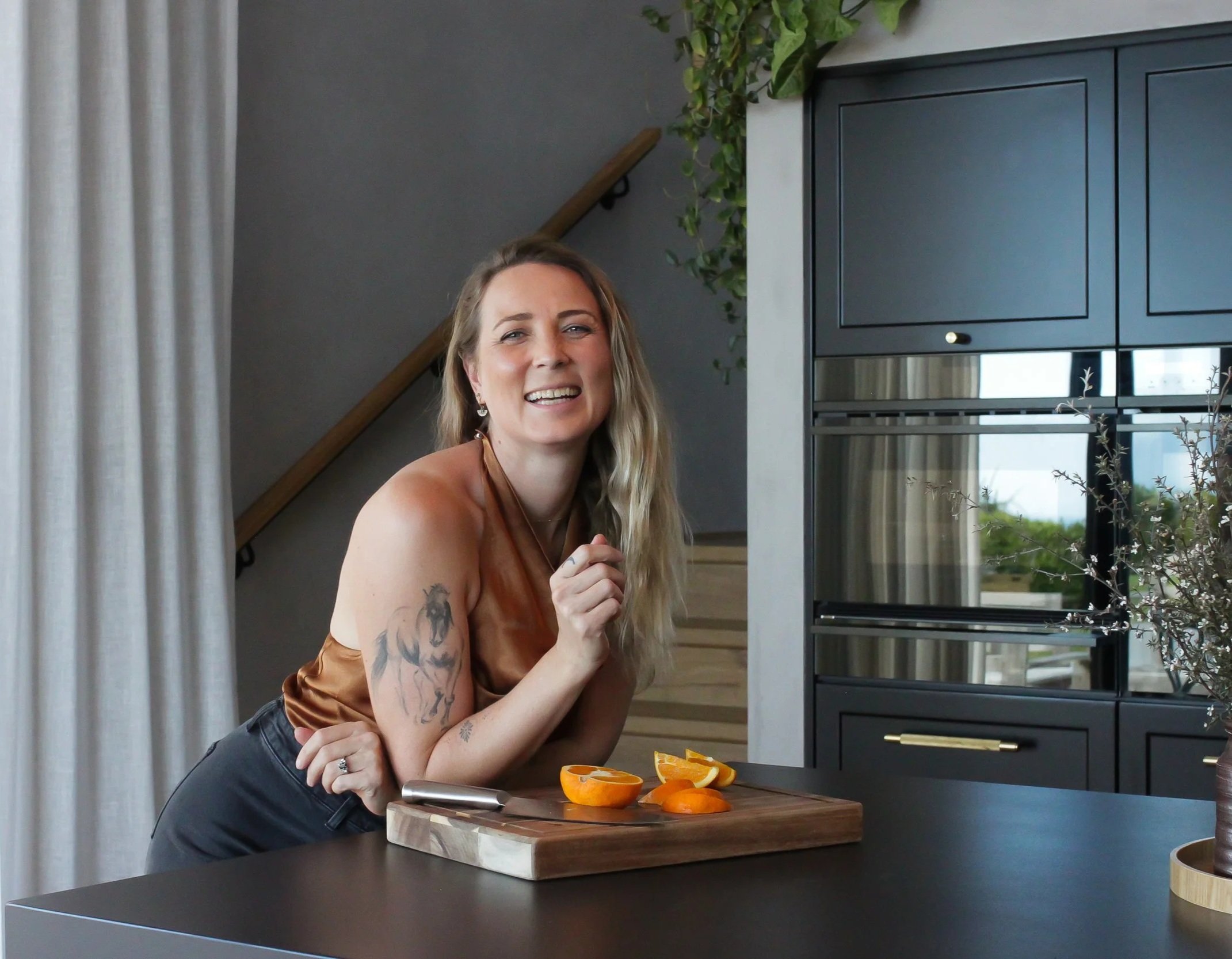 A woman with tattoos on her arm, smiling in a modern kitchen, leaning on a kitchen island with sliced oranges on a wooden cutting board.
