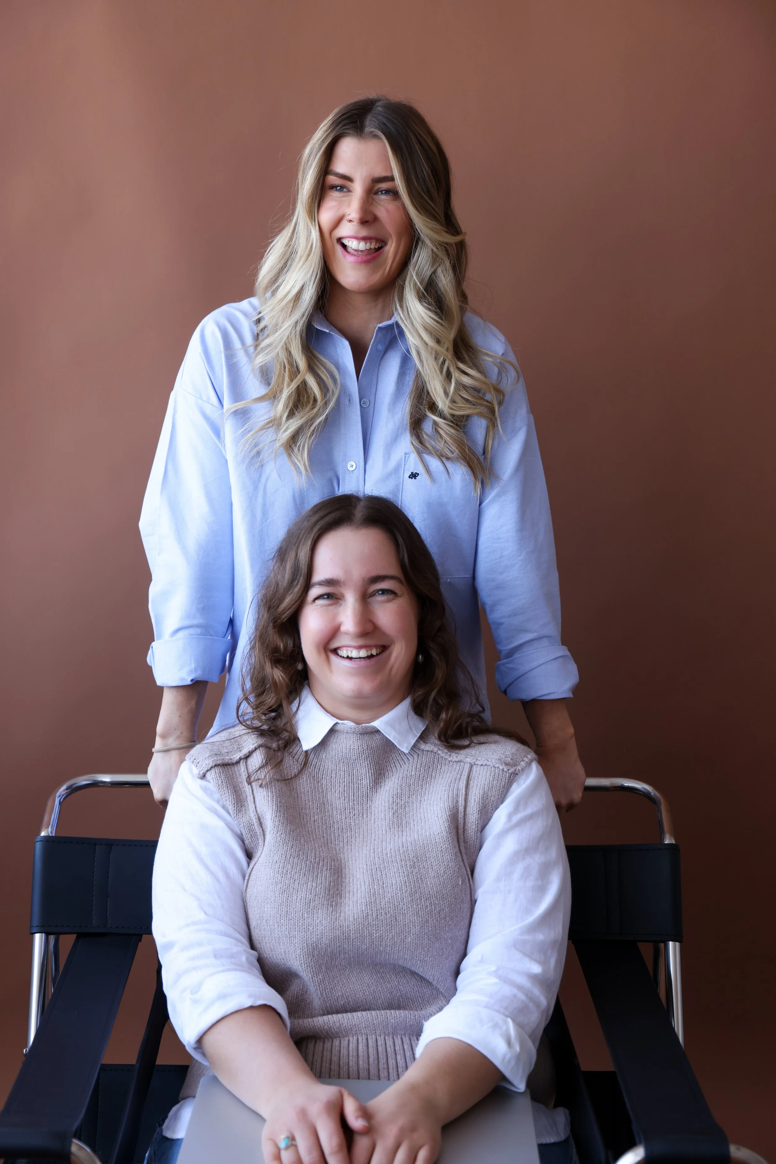 Two women, one standing behind the other in a wheelchair, both smiling and looking happy, in front of a plain brown background.