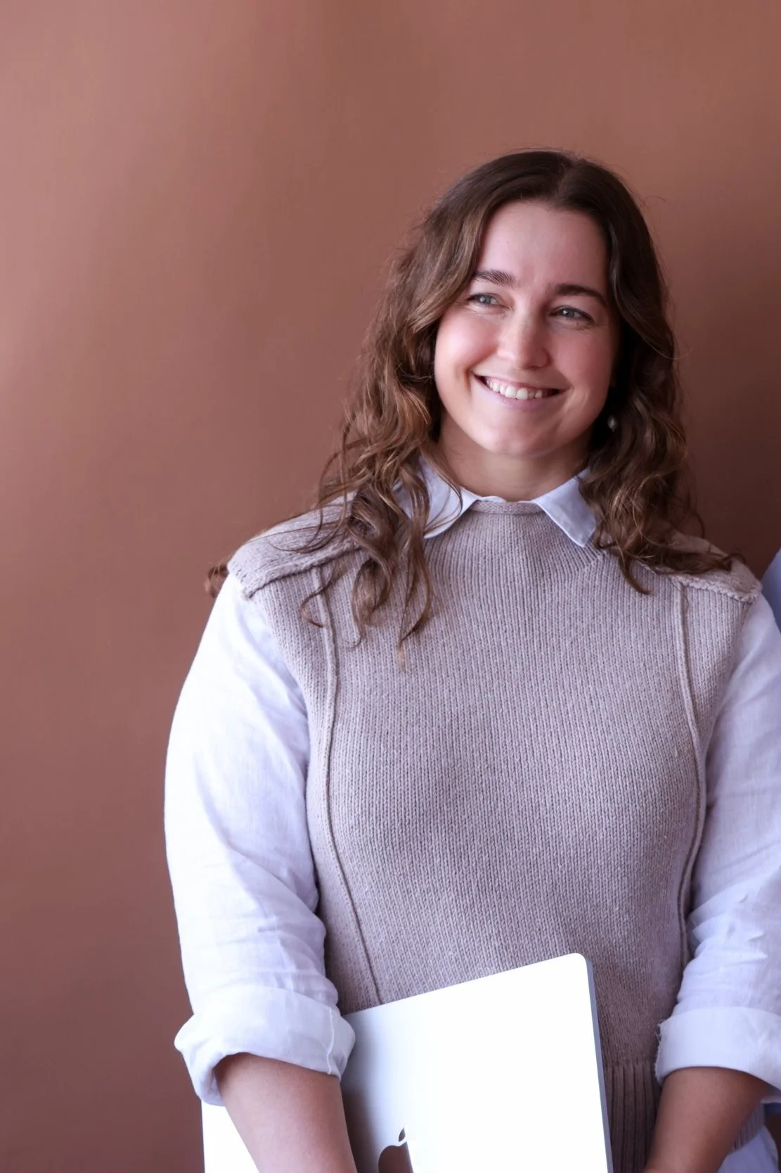A smiling woman with wavy brown hair wearing a white shirt and beige sweater vest, holding a silver MacBook, standing in front of a plain brown background.