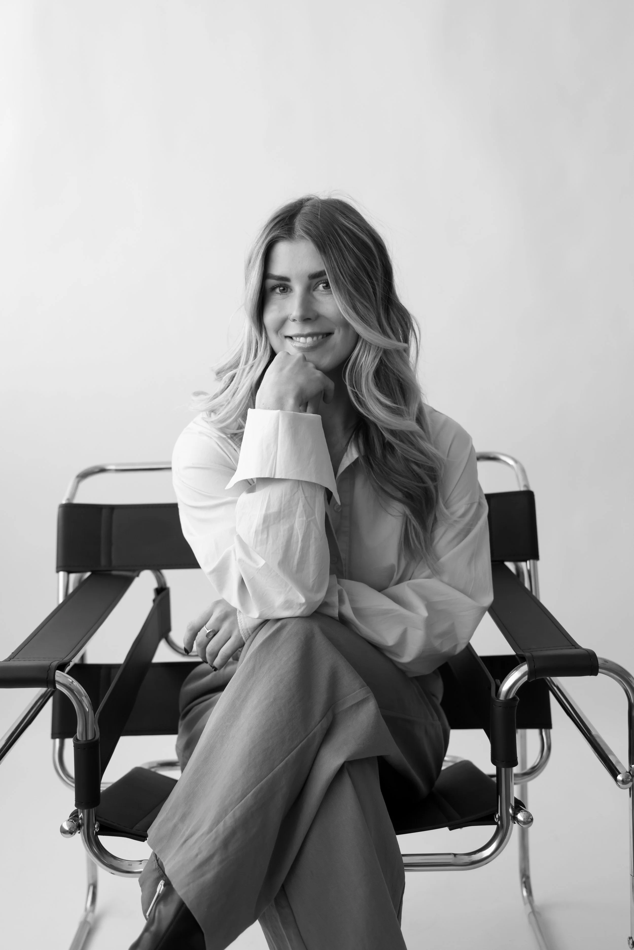 Black and white photo of a young woman sitting in a chair with crossed legs, smiling, wearing a light-colored blouse and dark pants, with her chin resting on her hand.