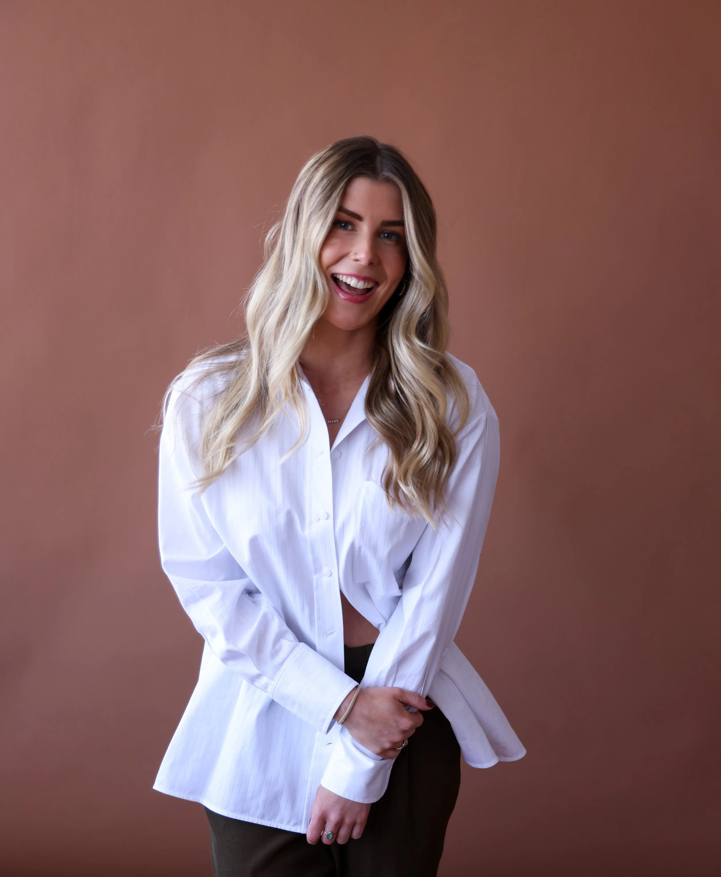 A young woman with long, wavy blonde hair, smiling and slightly tilting her head, wearing a white button-up shirt and dark pants, standing against a neutral brown background.