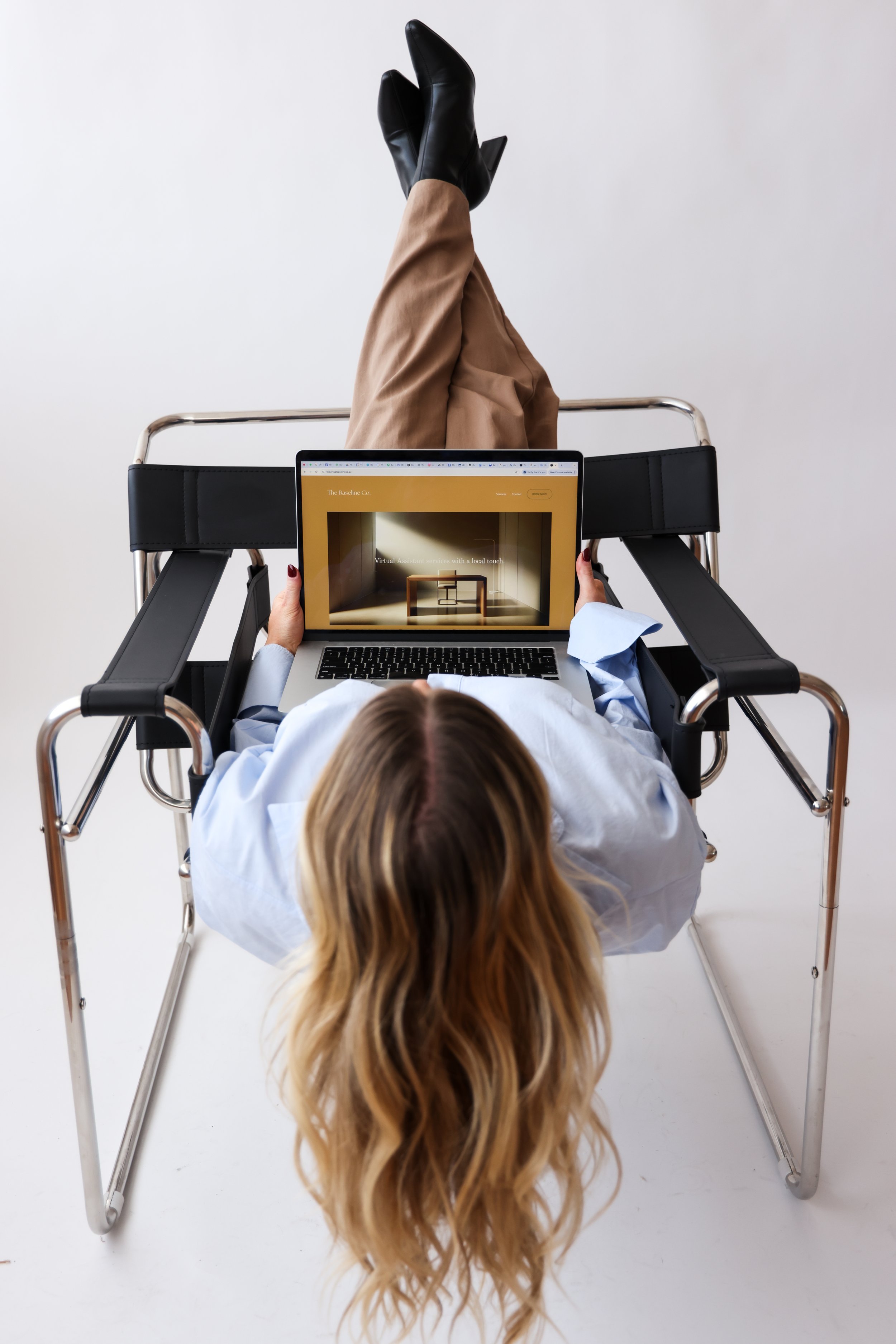 Woman lying on a hospital gurney using a laptop, with her legs raised and crossed, wearing black boots, tan pants, and a white shirt.