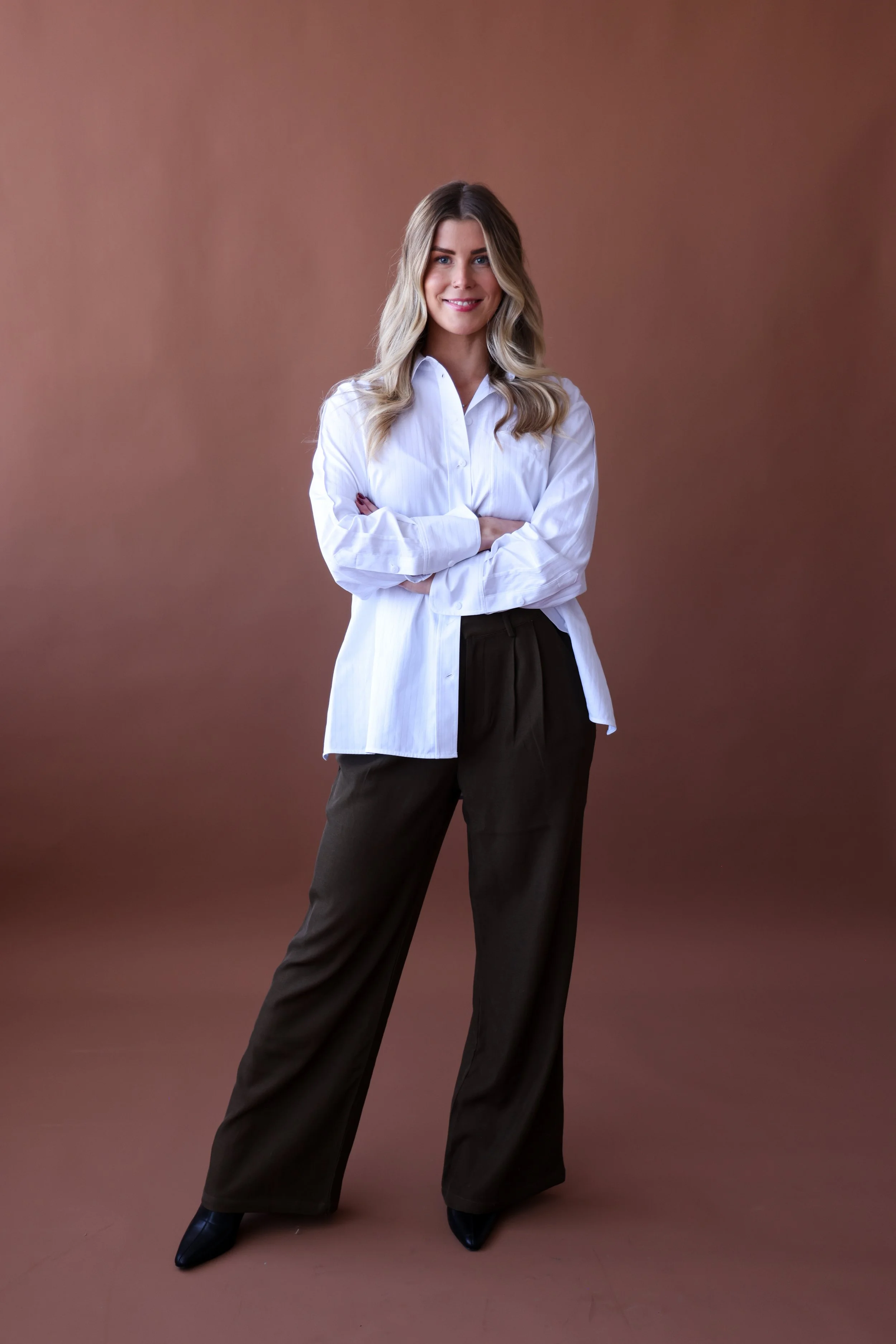 A woman standing with her arms crossed, wearing a white button-up shirt and dark wide-leg pants, smiling at the camera against a brown background.