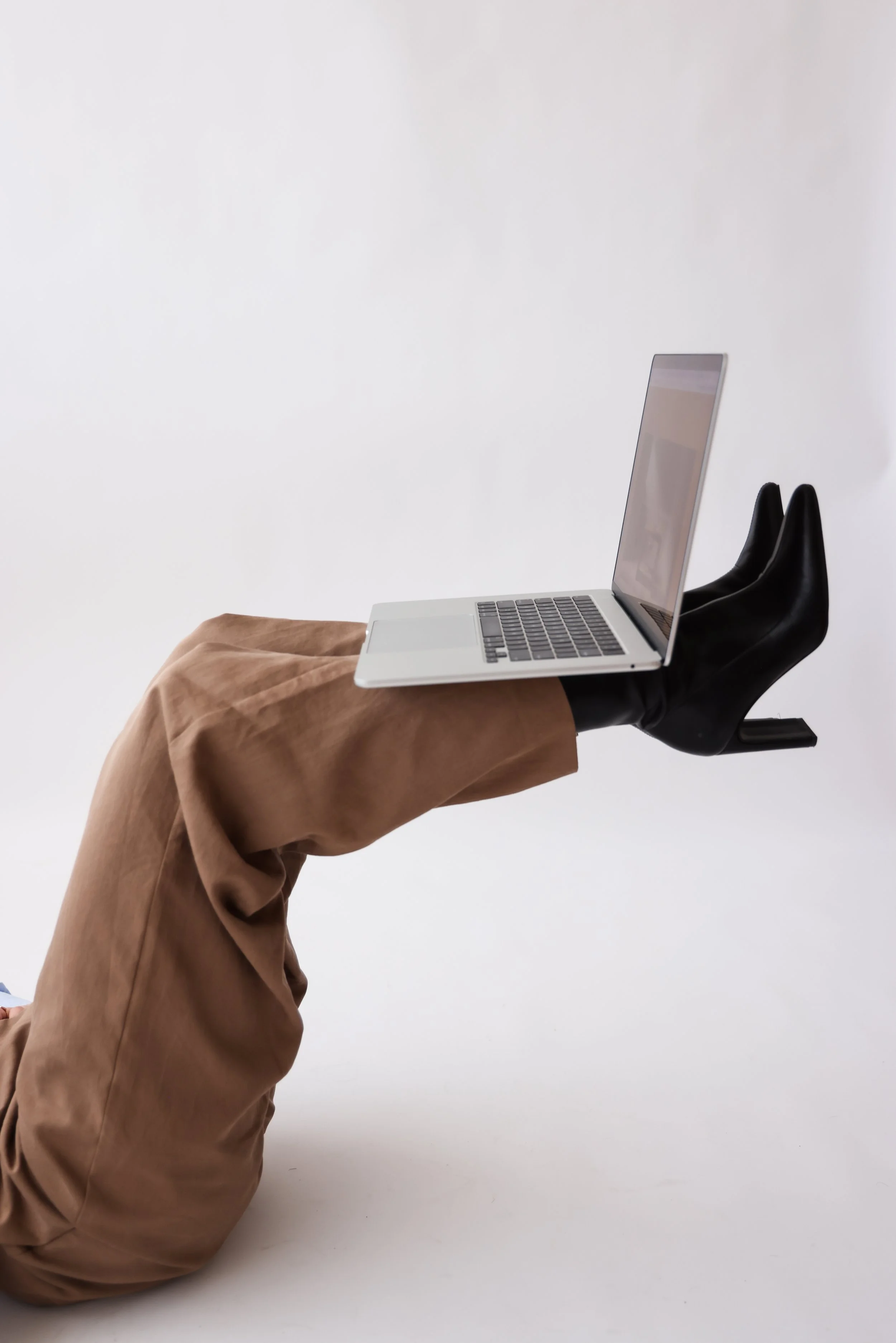 A person lying on the ground with legs raised, wearing brown pants and black heeled boots, holding a laptop on their feet against a plain white background.