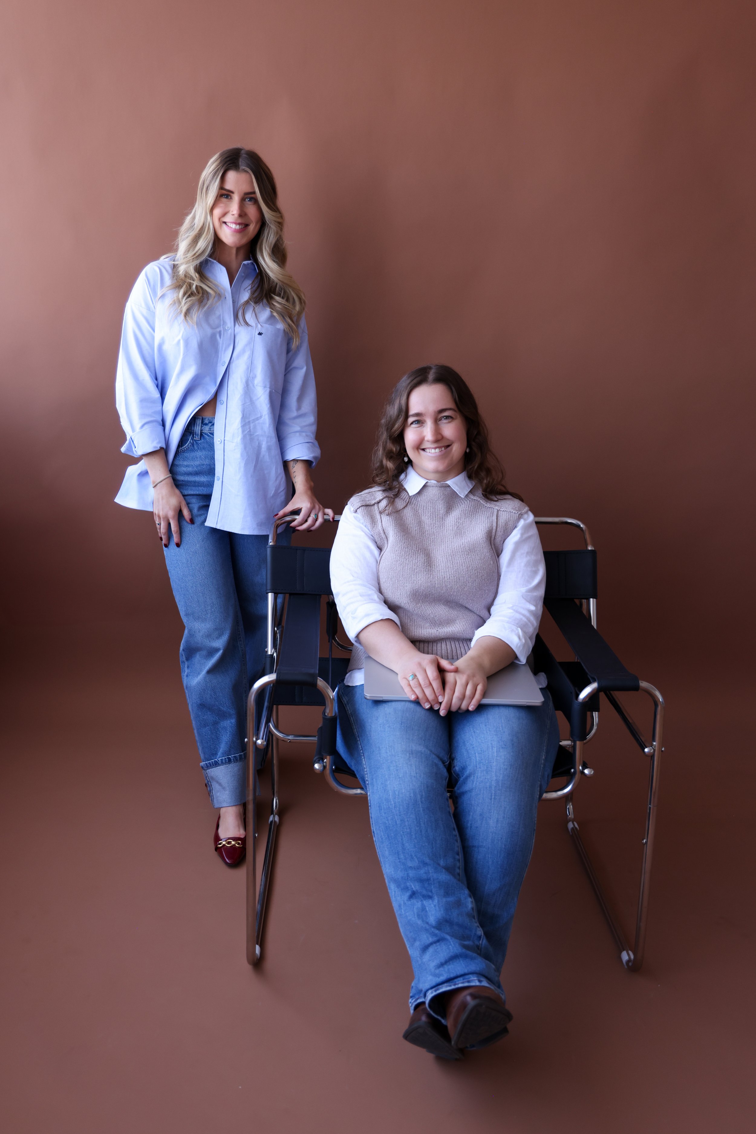 Two young women smiling, one standing in casual clothes and the other sitting in a wheelchair holding a laptop, against a plain brown background.