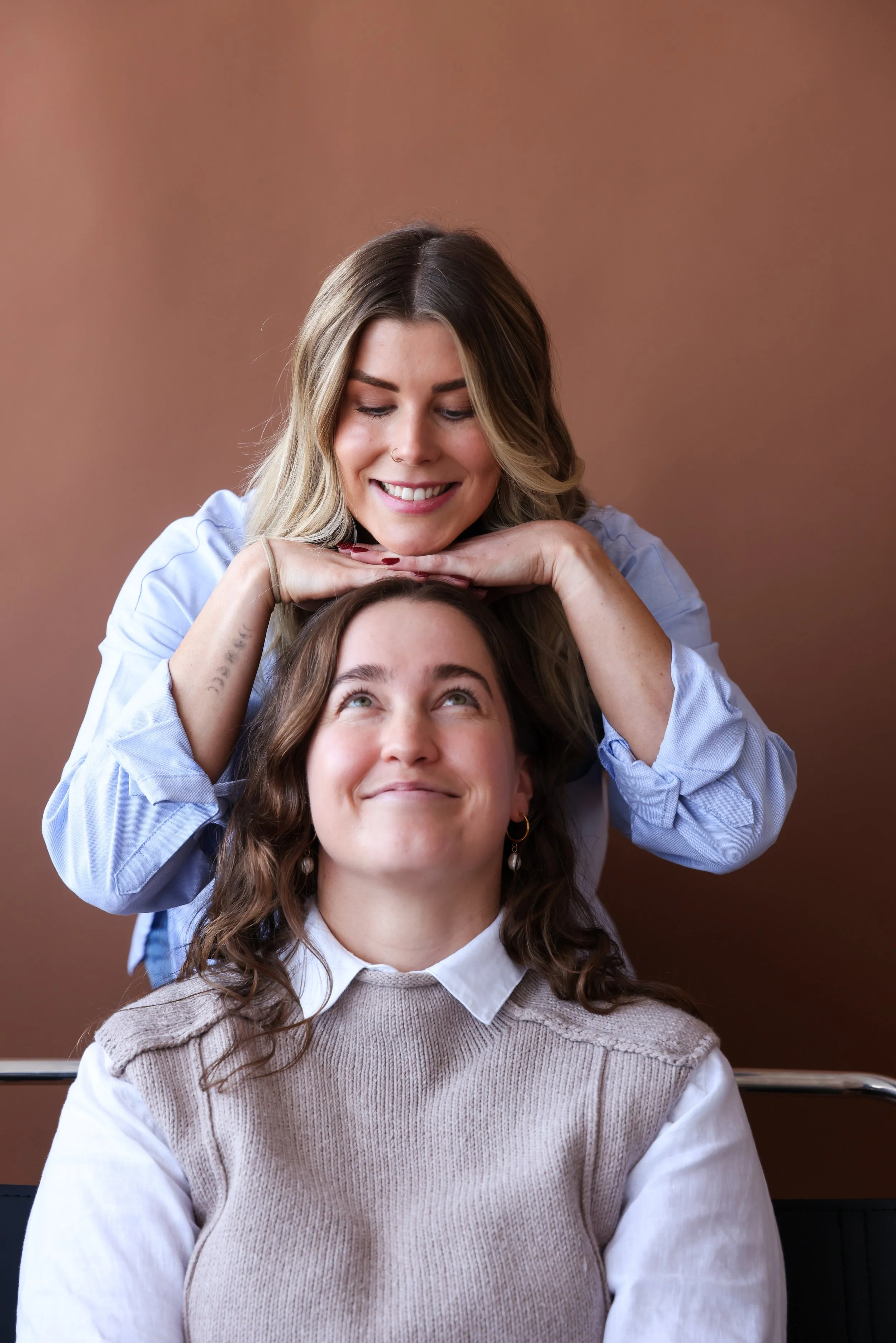Two women smiling and looking at each other, one sitting and the other leaning over her with her hands on the other's head, in a friendly pose against a brown background.