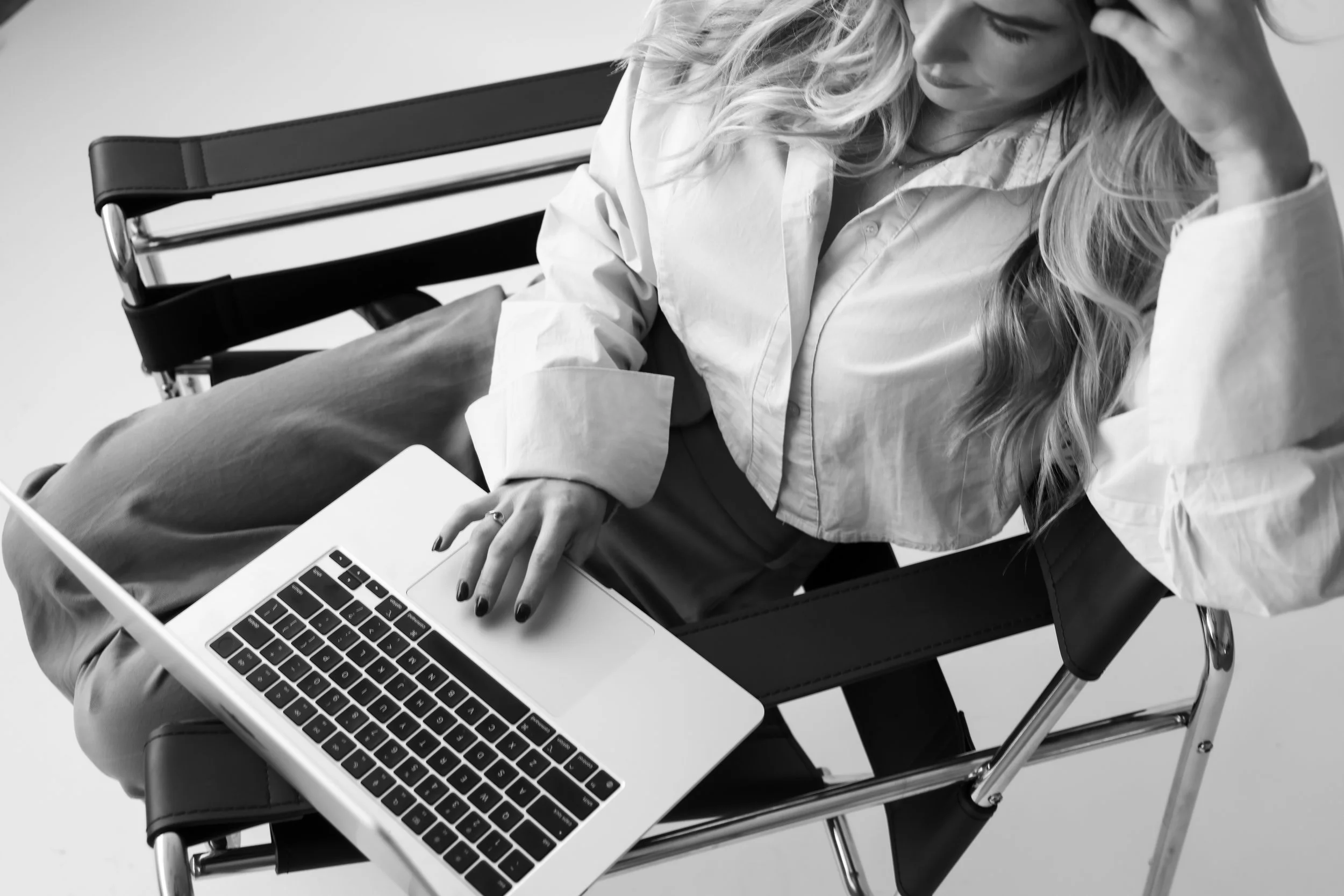 Black and white image of a woman sitting on a black medical or hospital bed with a silver frame, looking at a laptop on her lap, wearing a loose button-up shirt and beige pants.