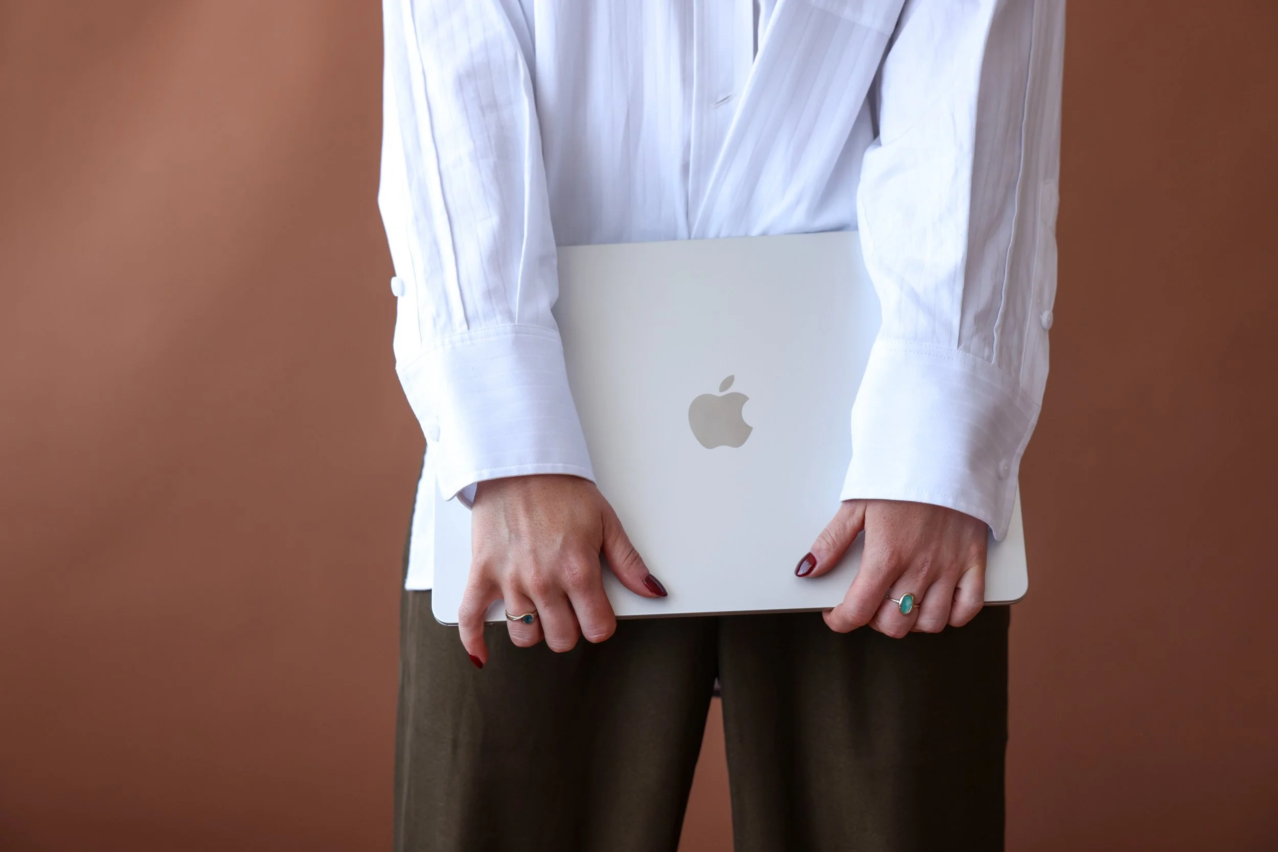 Person holding a silver MacBook with an Apple logo, wearing a white long-sleeve shirt and brown pants.
