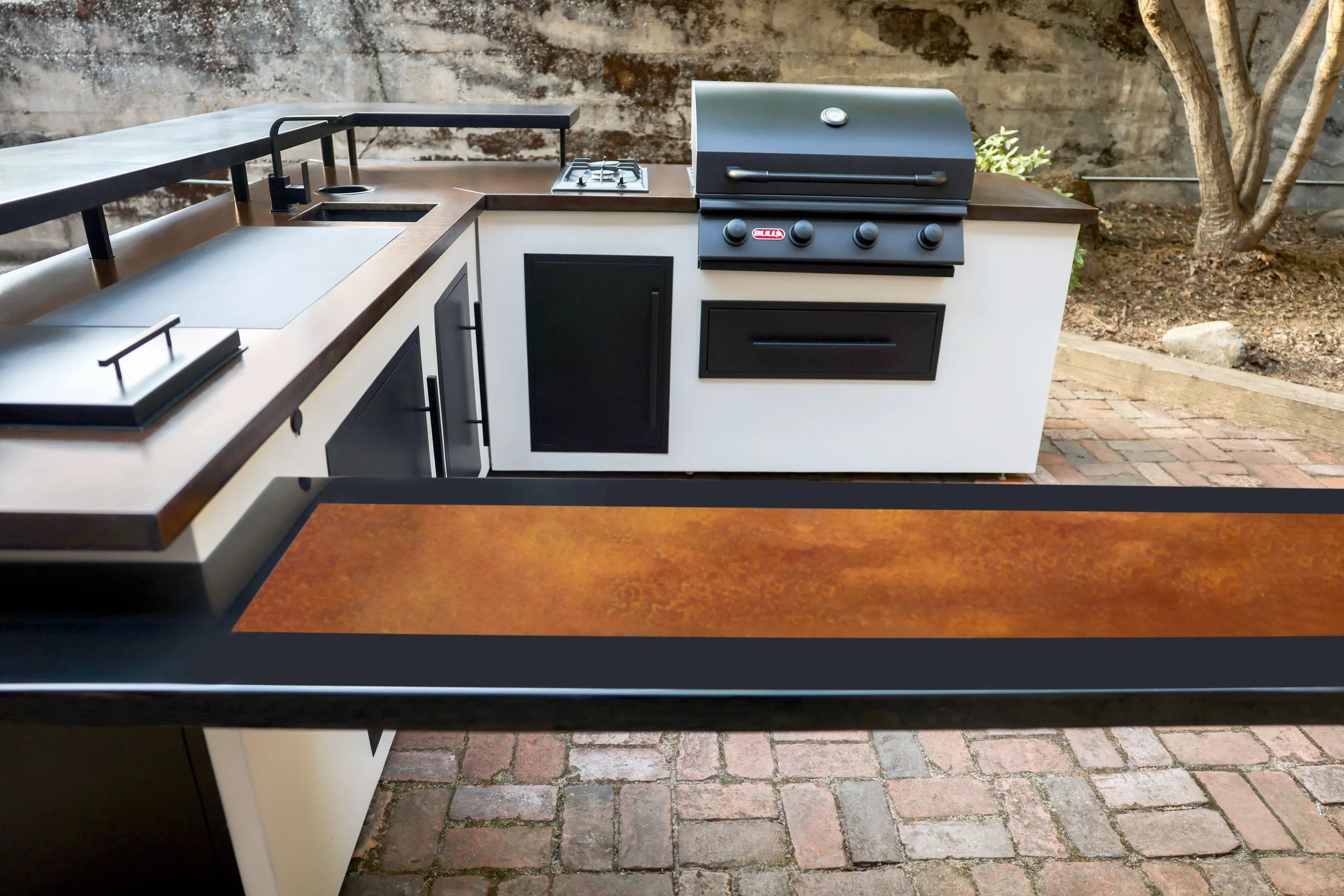 Outdoor kitchen with built-in grill, sink, and countertop on brick patio, with trees and a stone wall in the background.