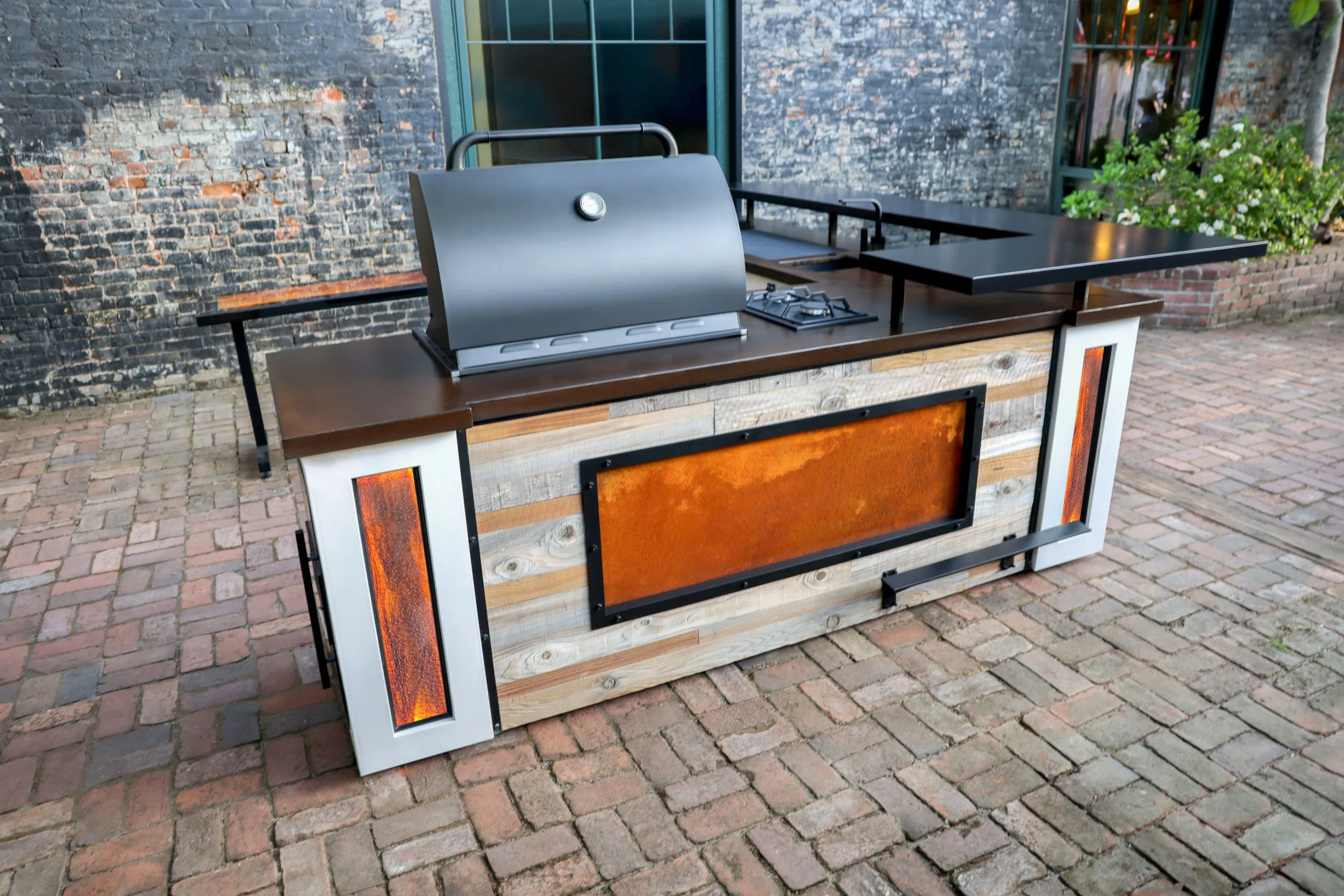 Outdoor kitchen with built-in grill, black stovetop, and rusty metal accents on a brick patio.