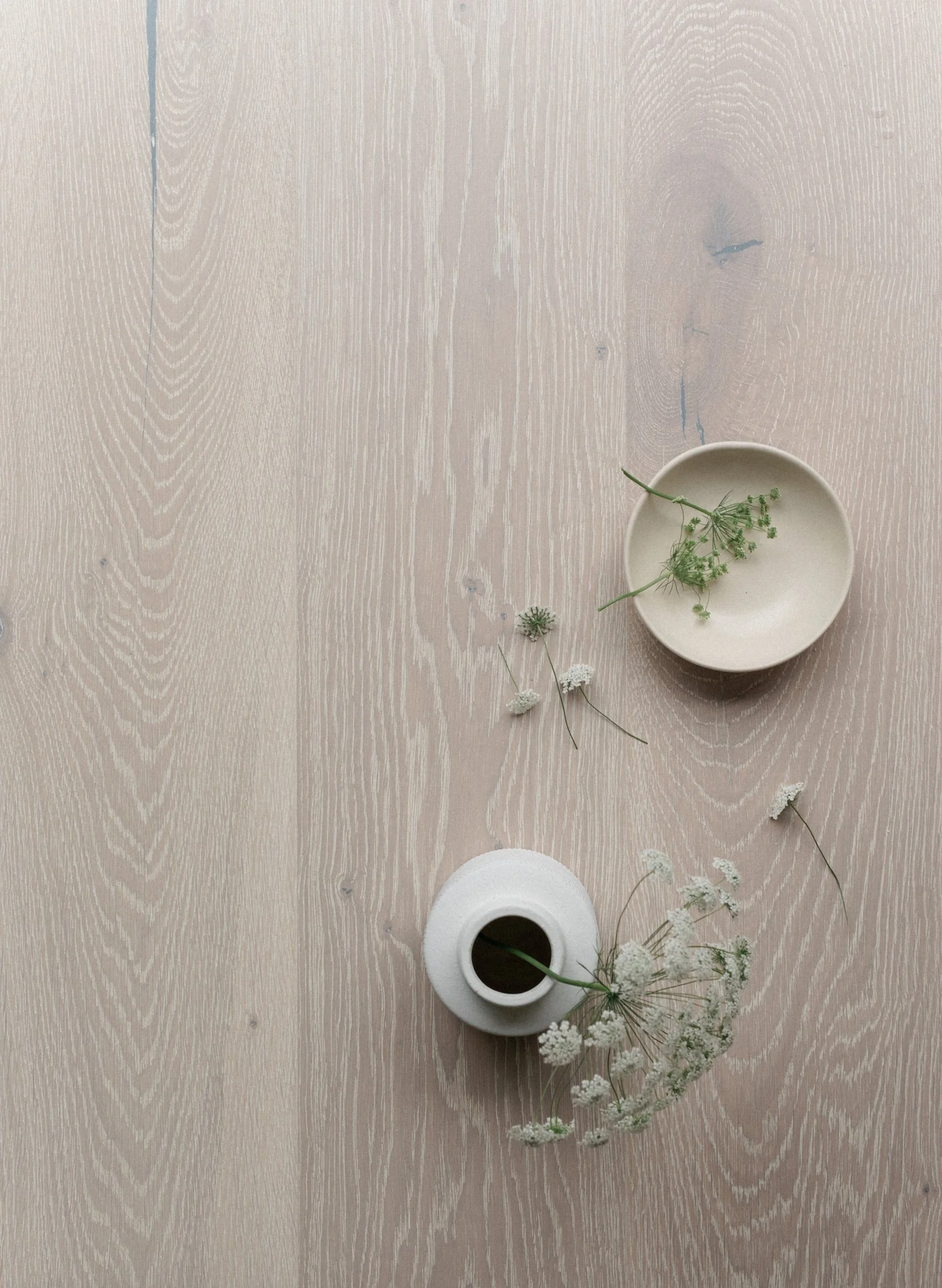 A top-down view of a light wood surface with a small beige ceramic bowl containing green sprigs, and a white vase with a narrow neck, holding white flowers, with some flowers and sprigs scattered around.