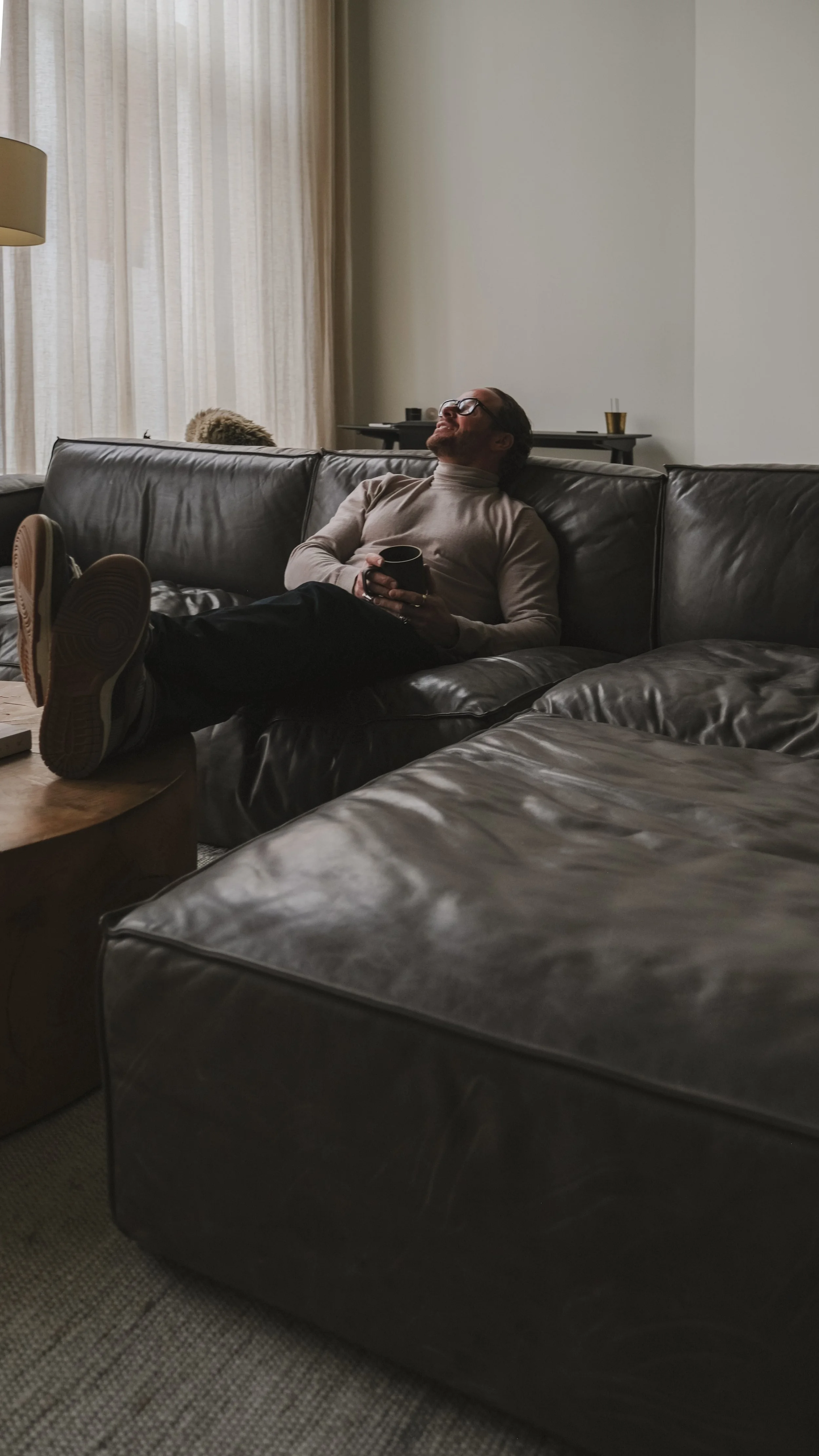 A man with glasses, wearing a turtleneck, is lounging on a dark leather sofa with his feet resting on a coffee table, holding a mug, and smiling in a cozy living room.