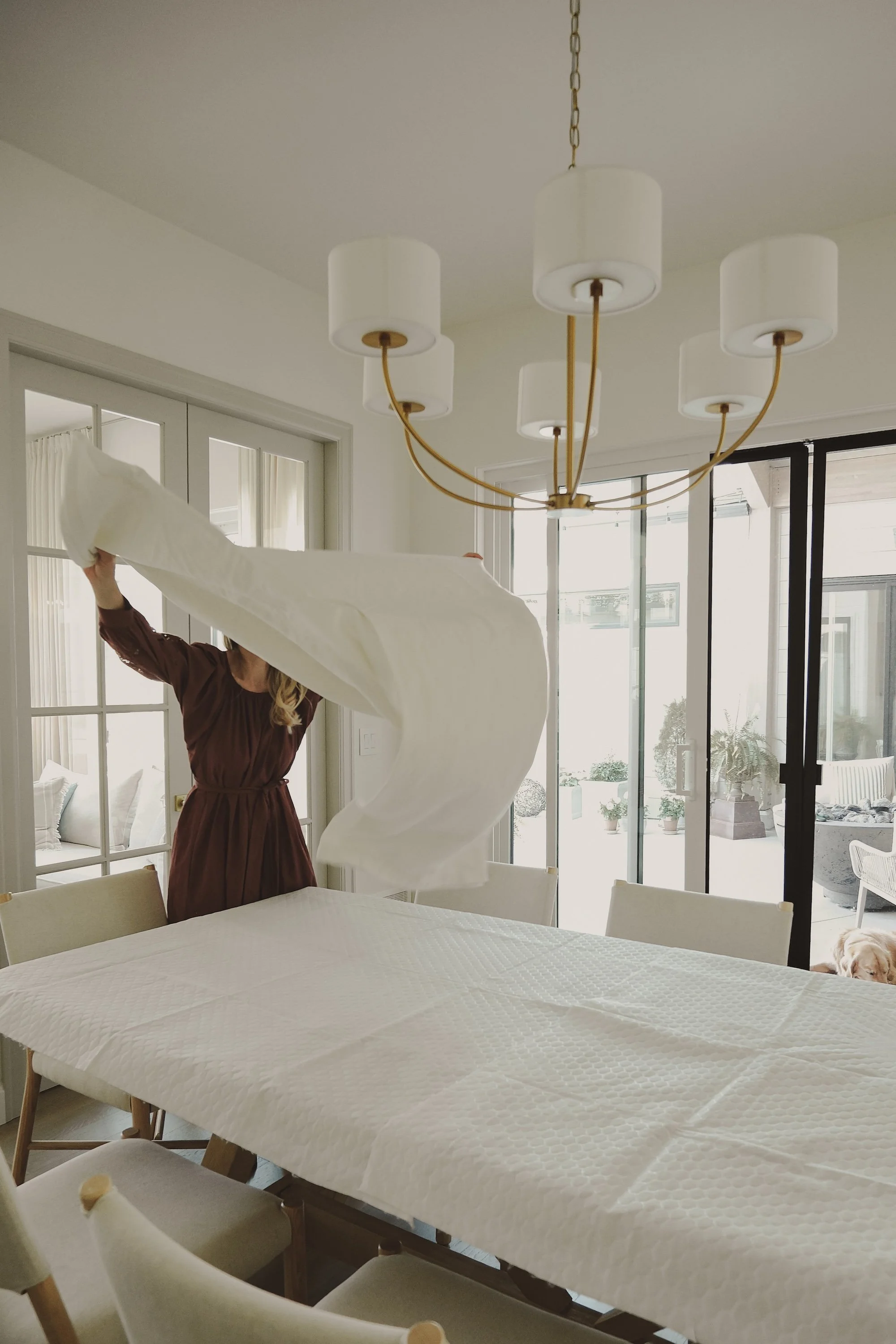 A woman in a brown dress is tossing a white bed sheet inside a dining room with a table, chairs, and large glass doors leading to an outdoor patio.