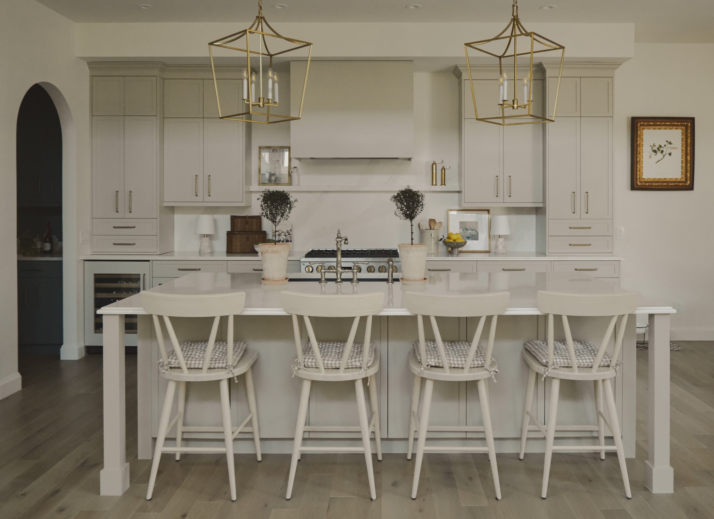 Modern kitchen with white cabinetry, a central island with four white chairs, two potted trees, pendant light fixtures, and decorative wall art.
