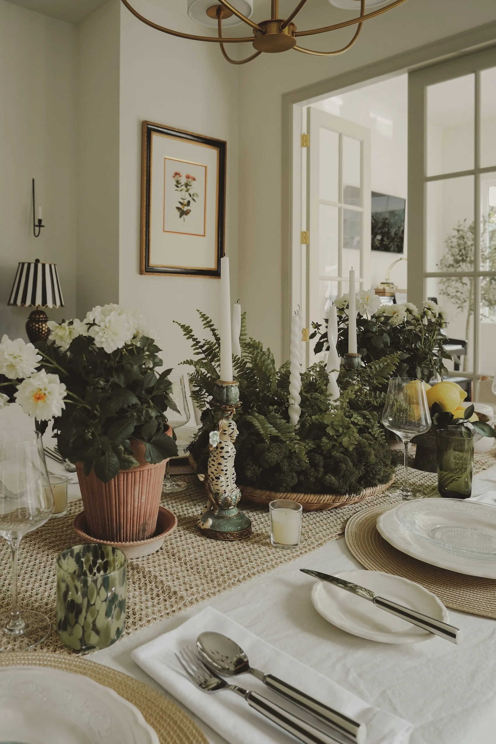 A dining table set with plates, utensils, glasses, and candles, decorated with floral arrangements and greenery, inside a bright room with a door opening to another room.