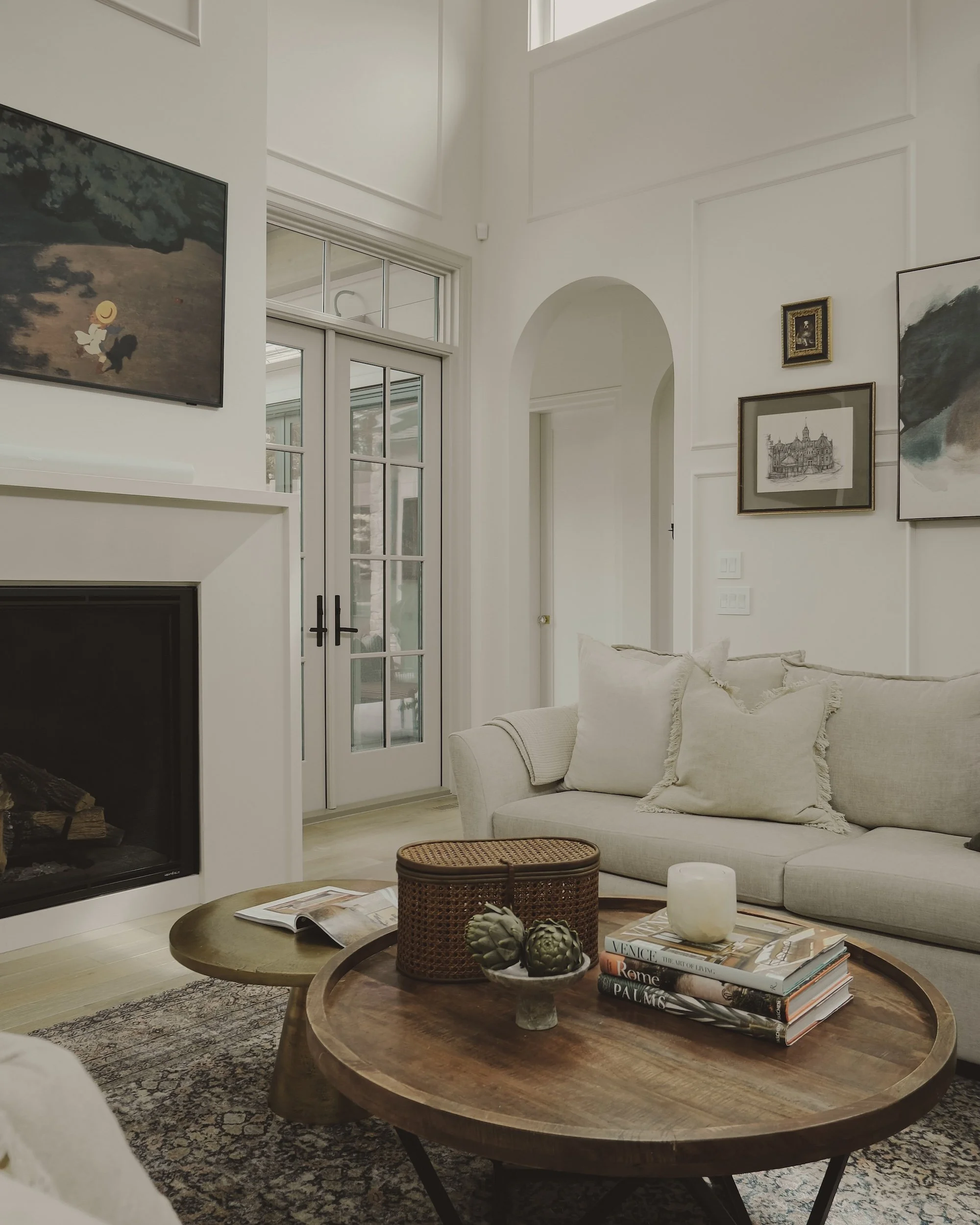 Bright living room with white walls and art, beige sofa with cushions, wooden coffee and side tables with books, decorative artichokes, candle, fireplace, and french doors.