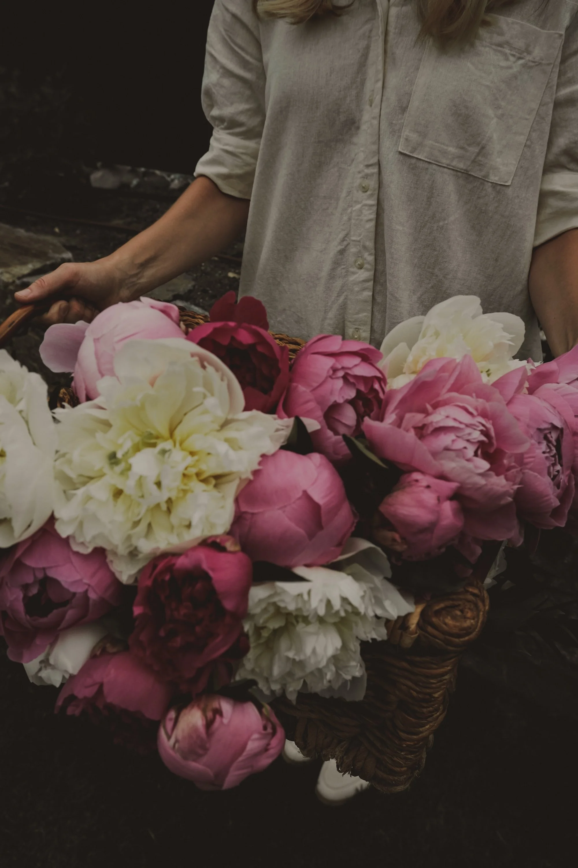Person holding a basket of pink, white, and red peonies flowers, wearing a beige shirt.