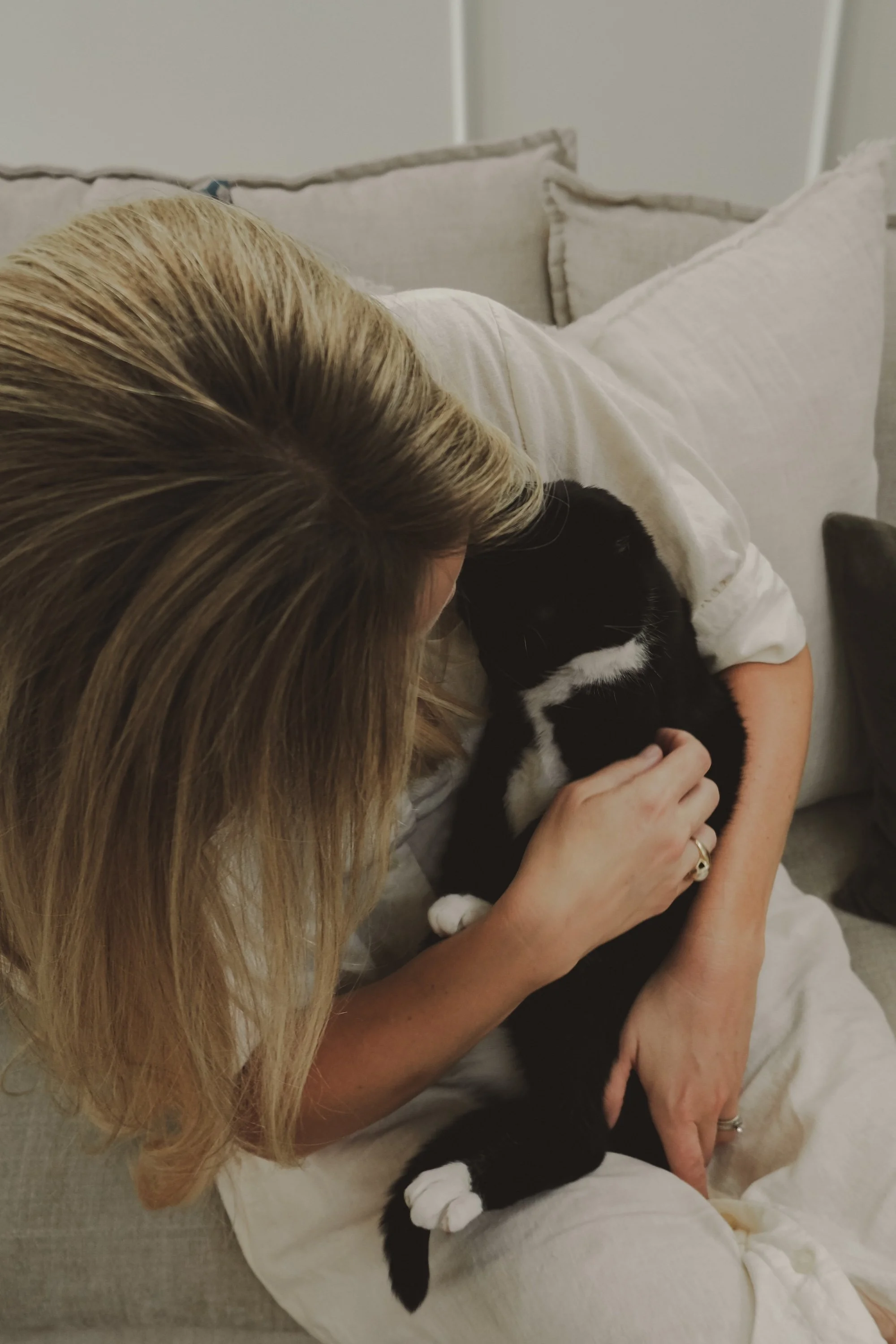 A woman with blonde hair lying on a beige couch, holding a black and white kitten close to her chest.