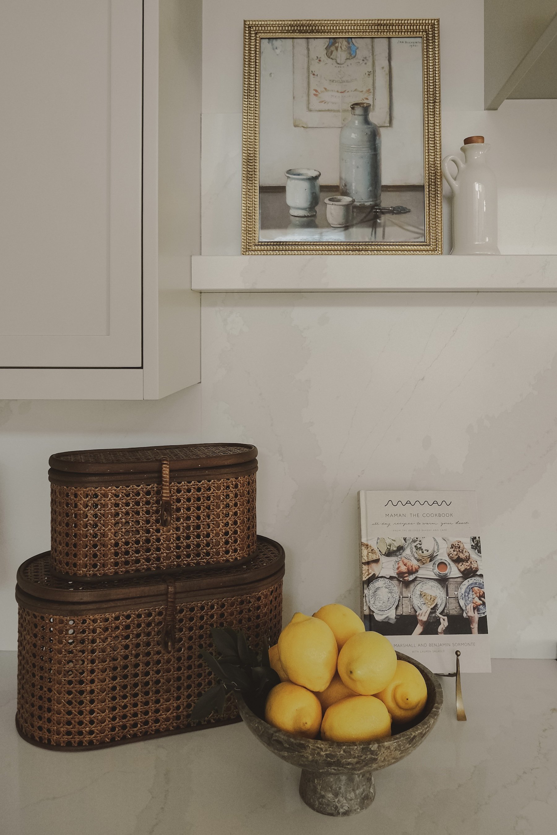 Decorative kitchen display with woven baskets, a bowl of lemons, a cookbook, framed artwork, and ceramic vases on a white countertop.