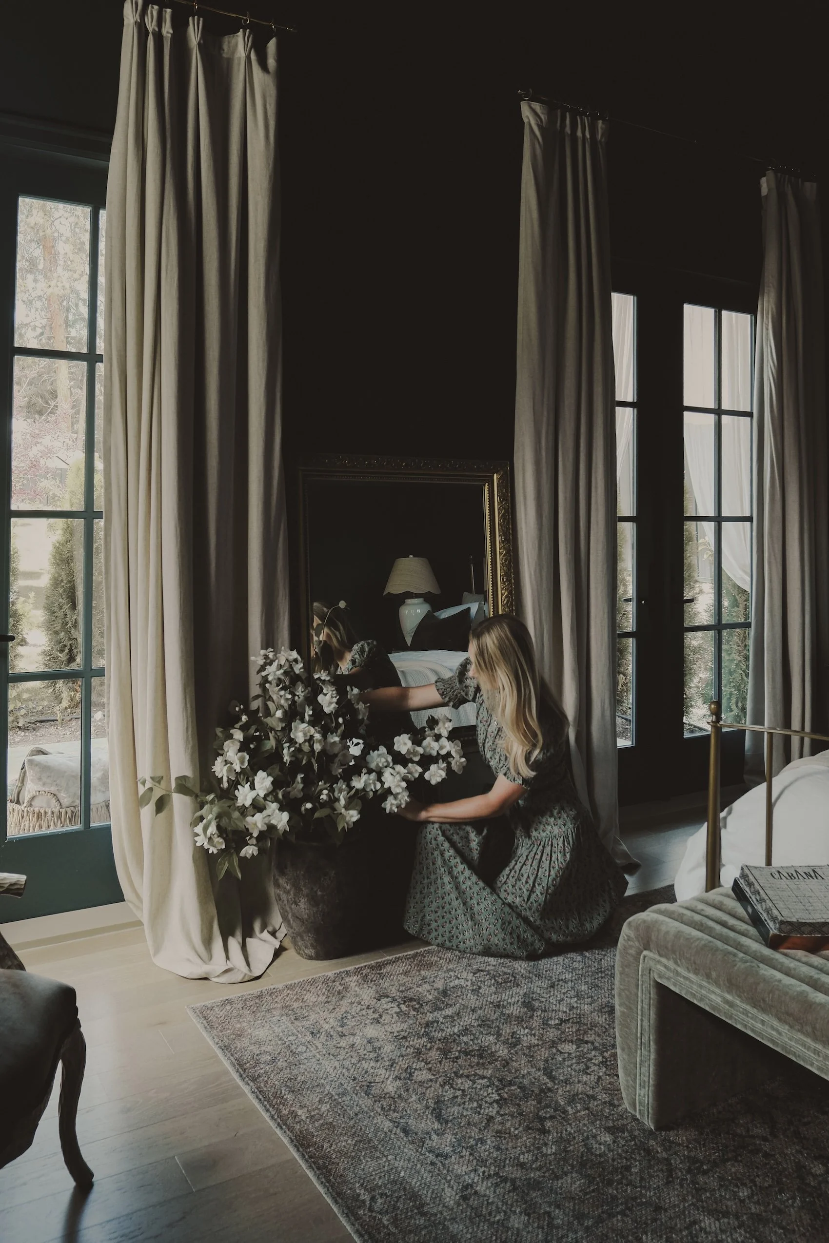 A woman with blonde hair kneeling on the floor, arranging white flowers in a vase next to a mirror in a living room with large windows and beige curtains.