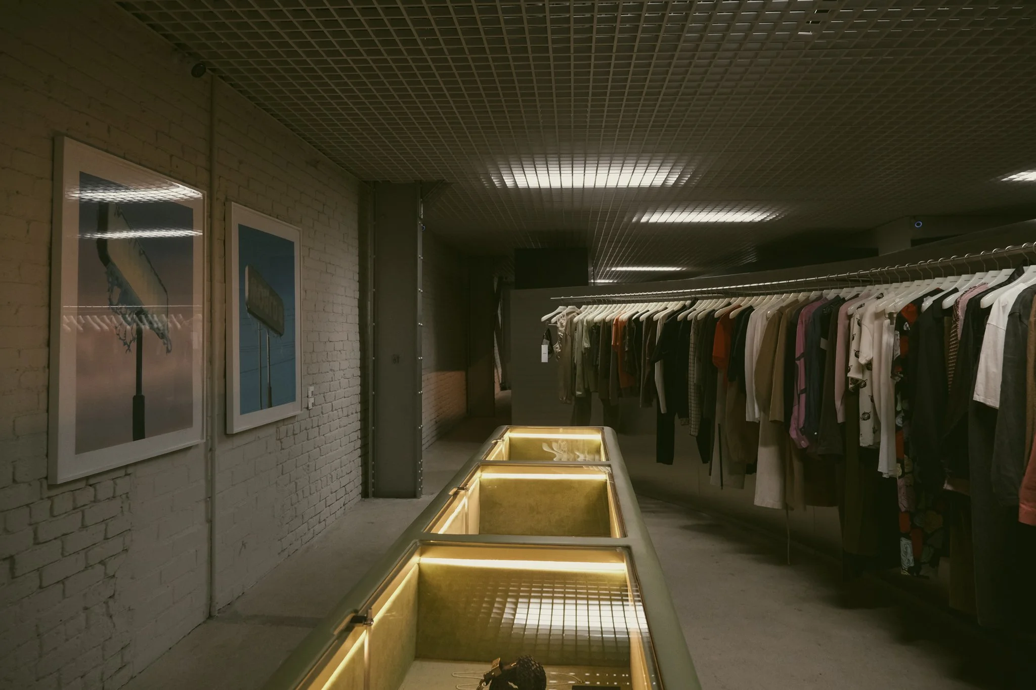 A clothing store with a rack of hanging shirts and jackets on the right side, lit display cases in the center, and framed pictures of signs on the brick wall to the left.