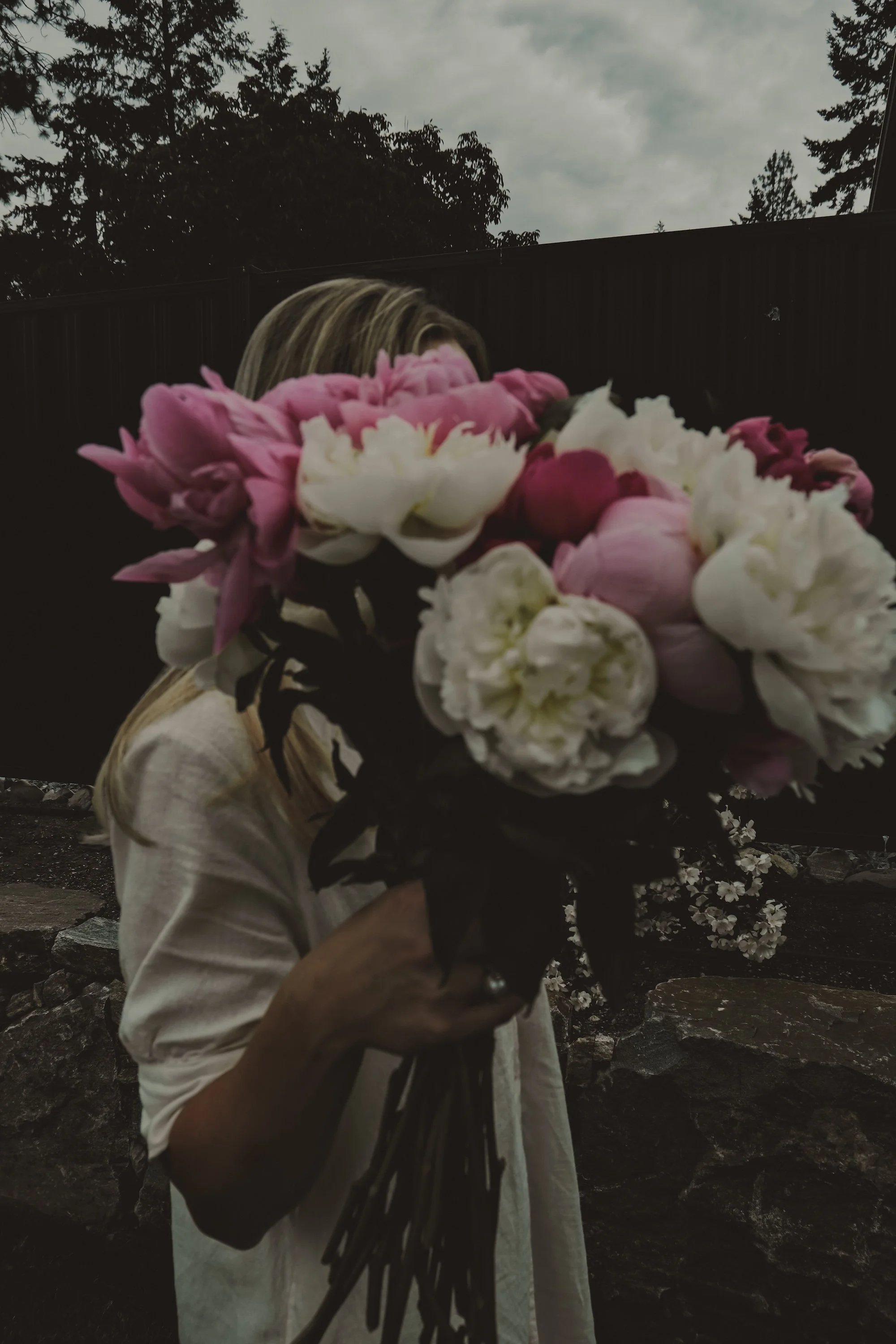 A person holding a large bouquet of pink and white peonies, with their face hidden behind the flowers. The background shows an outdoor setting with trees and a cloudy sky.