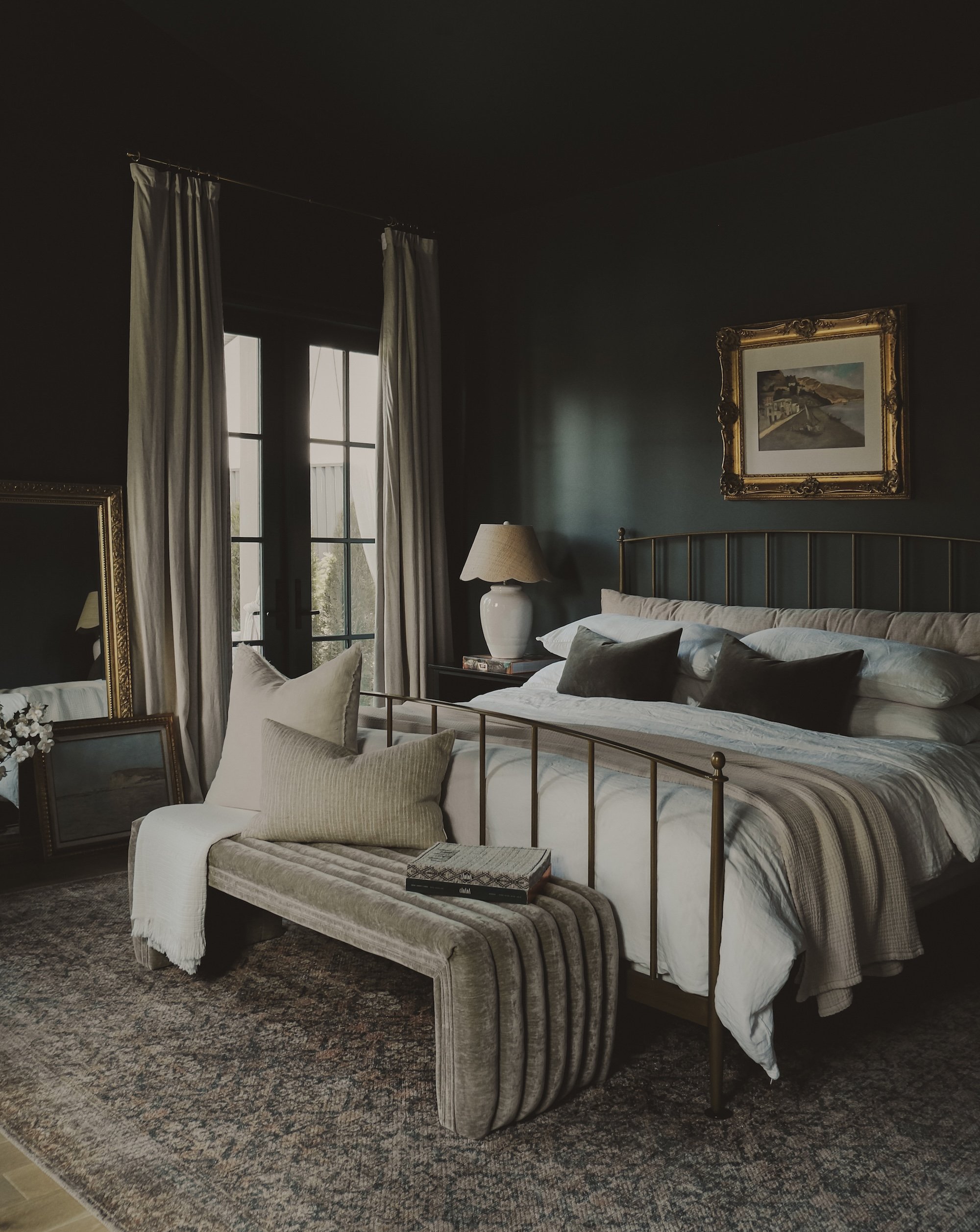 A cozy bedroom with a brass bed, white bedding, and dark pillows. A side table with a lamp, a mirror, and a framed picture on the wall. A window with white curtains and a patterned bench at the foot of the bed.