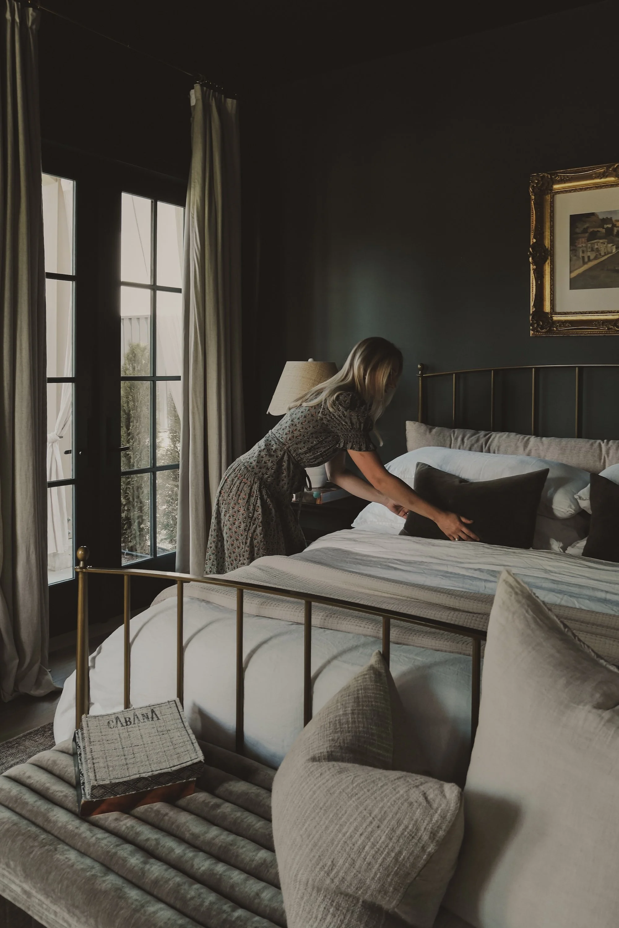 A woman in a patterned dress making the bed in a dark-colored bedroom with beige curtains, a gold-framed painting, and a brass bedframe.