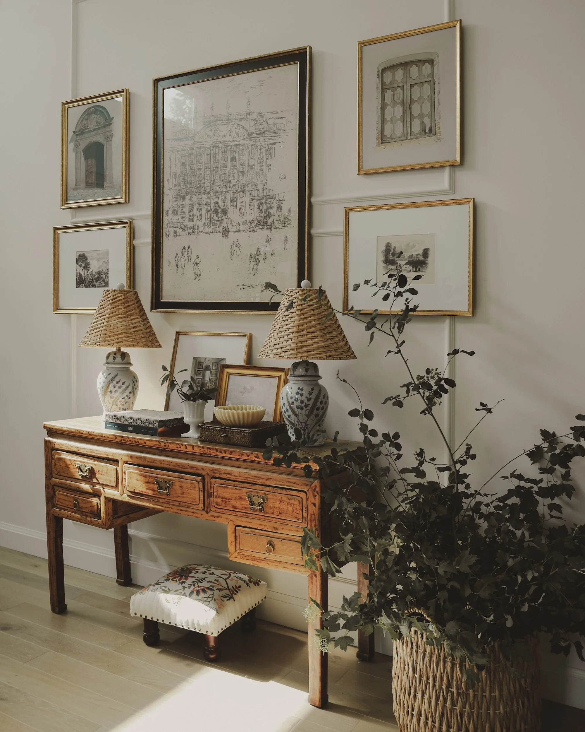 A vintage wooden console table with two lamps, decorative vases, pictures, and a bowl, positioned against a wall with six framed pictures and a large potted leafy plant nearby.