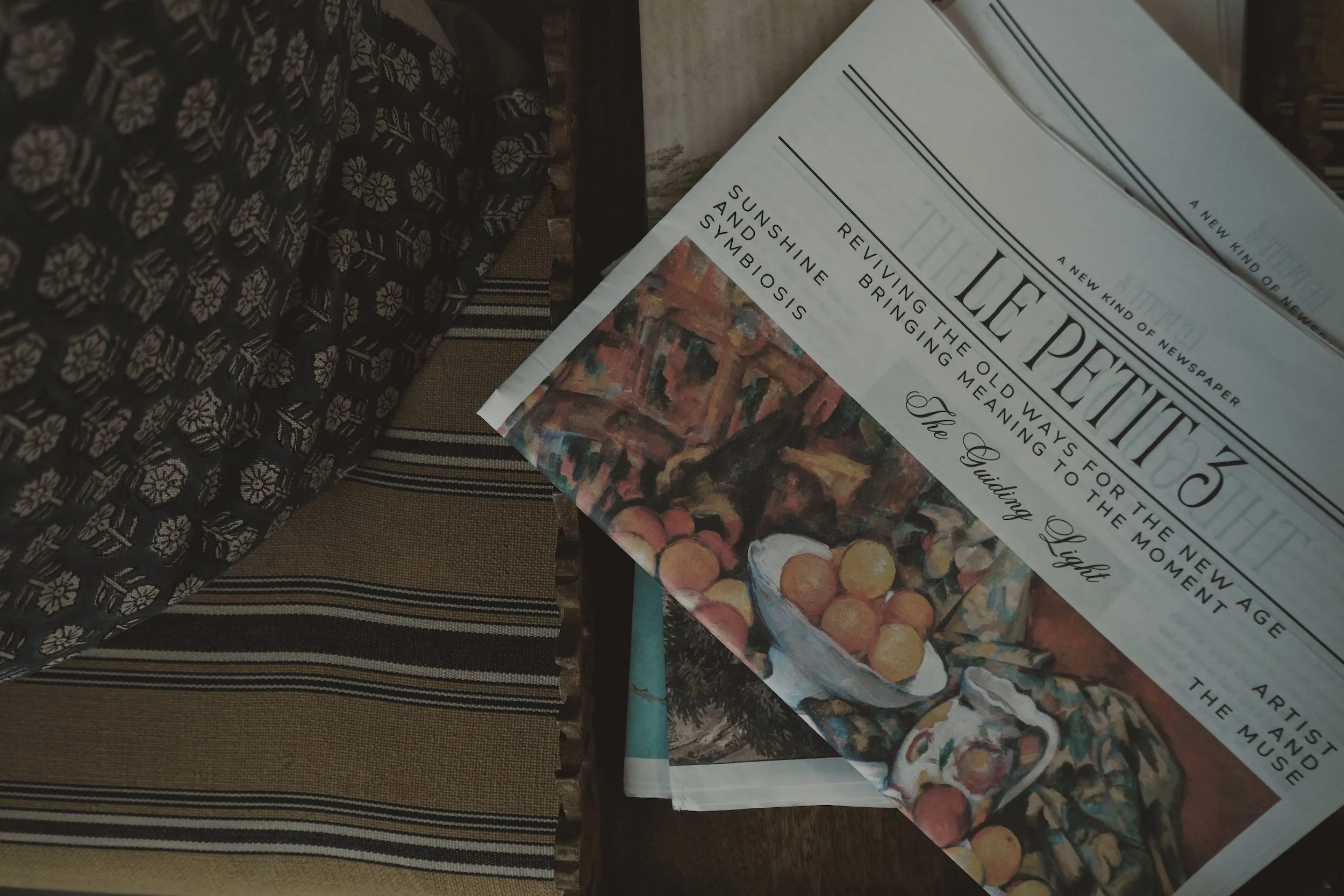 A stack of newspapers on a table, with the top one titled 'The Lee Petition.' The newspaper features a painting of a bowl of oranges and a pitcher, with headlines about art, old age, and the meaning of moments.