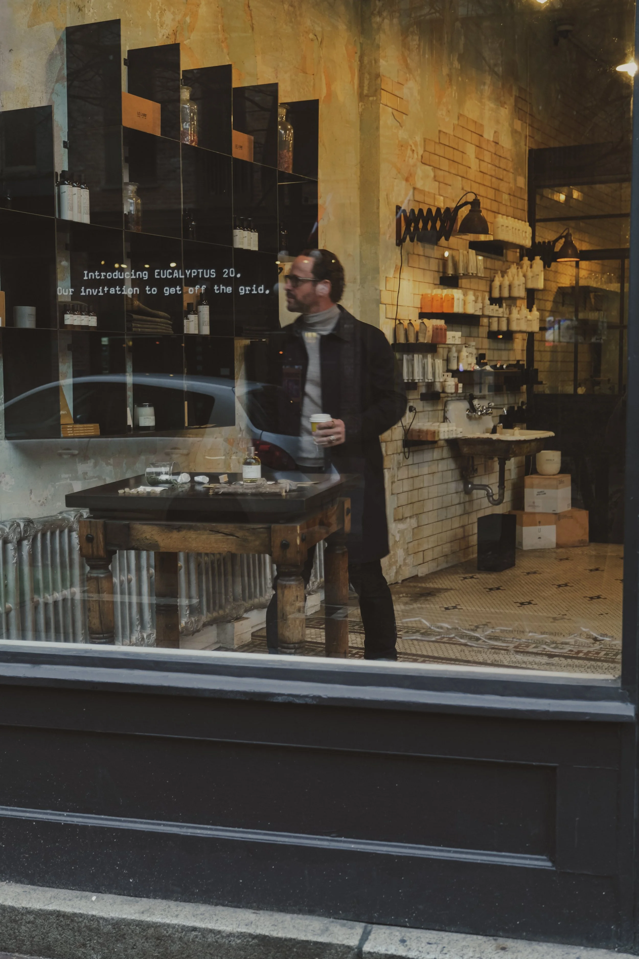A man inside a rustic shop or cafe, holding a coffee cup, looking to the side, with shelves of bottles and products on the wall behind him, and a tablet or sign displaying text about a new product.