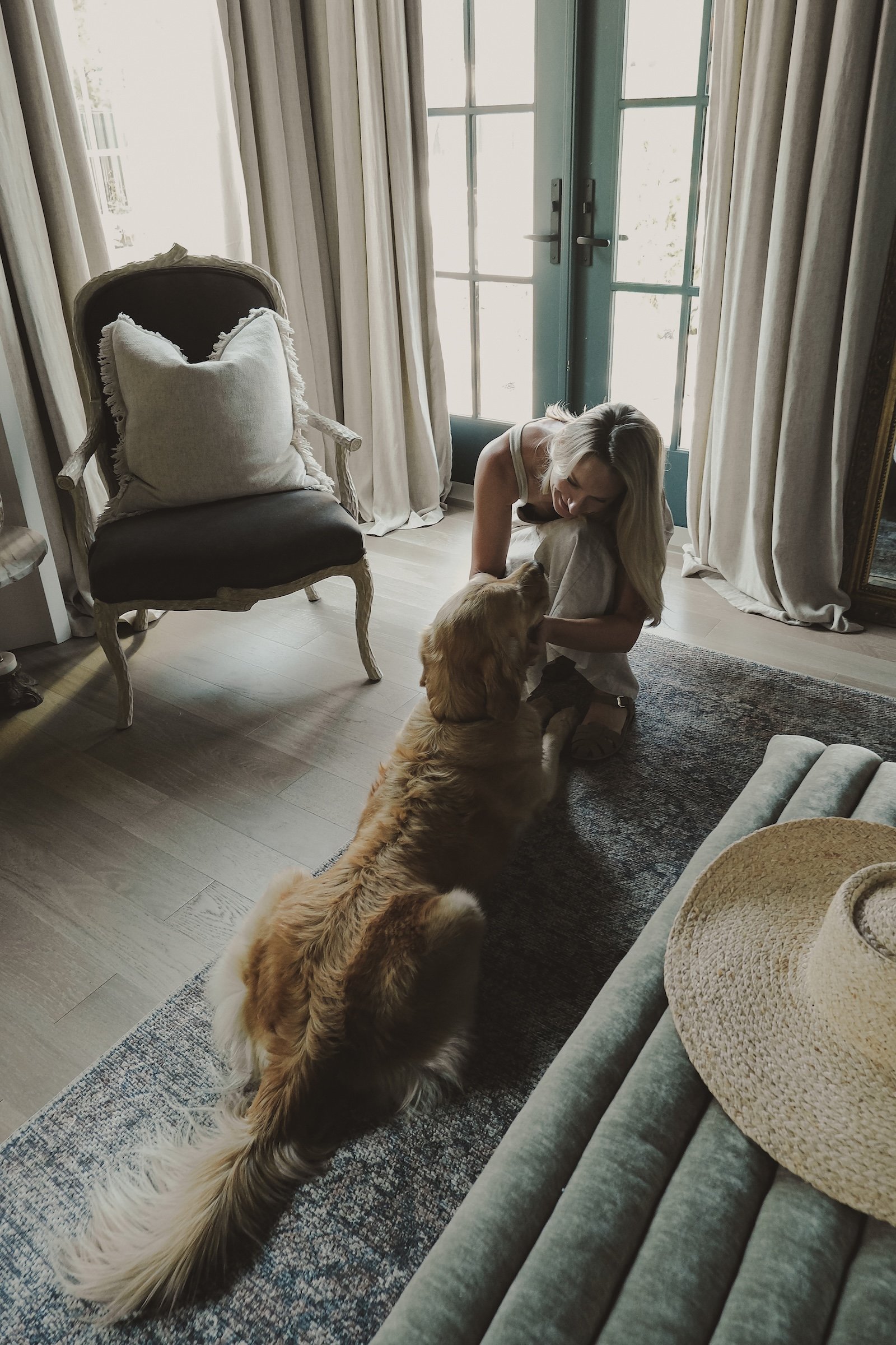 Woman kneeling on the floor with a golden retriever dog inside a living room with natural light.