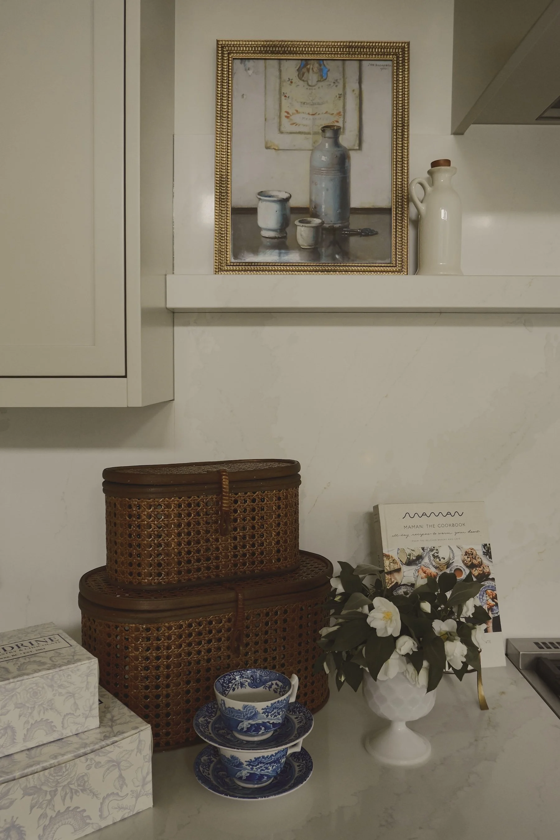 A kitchen counter with decorative items, including a white vase with white flowers, stacked blue and white dishes, bamboo containers, a cookbook titled 'MAMAN, THE COOKBOOK,' and a picture frame with a still life painting of bottles and cups on the wall.