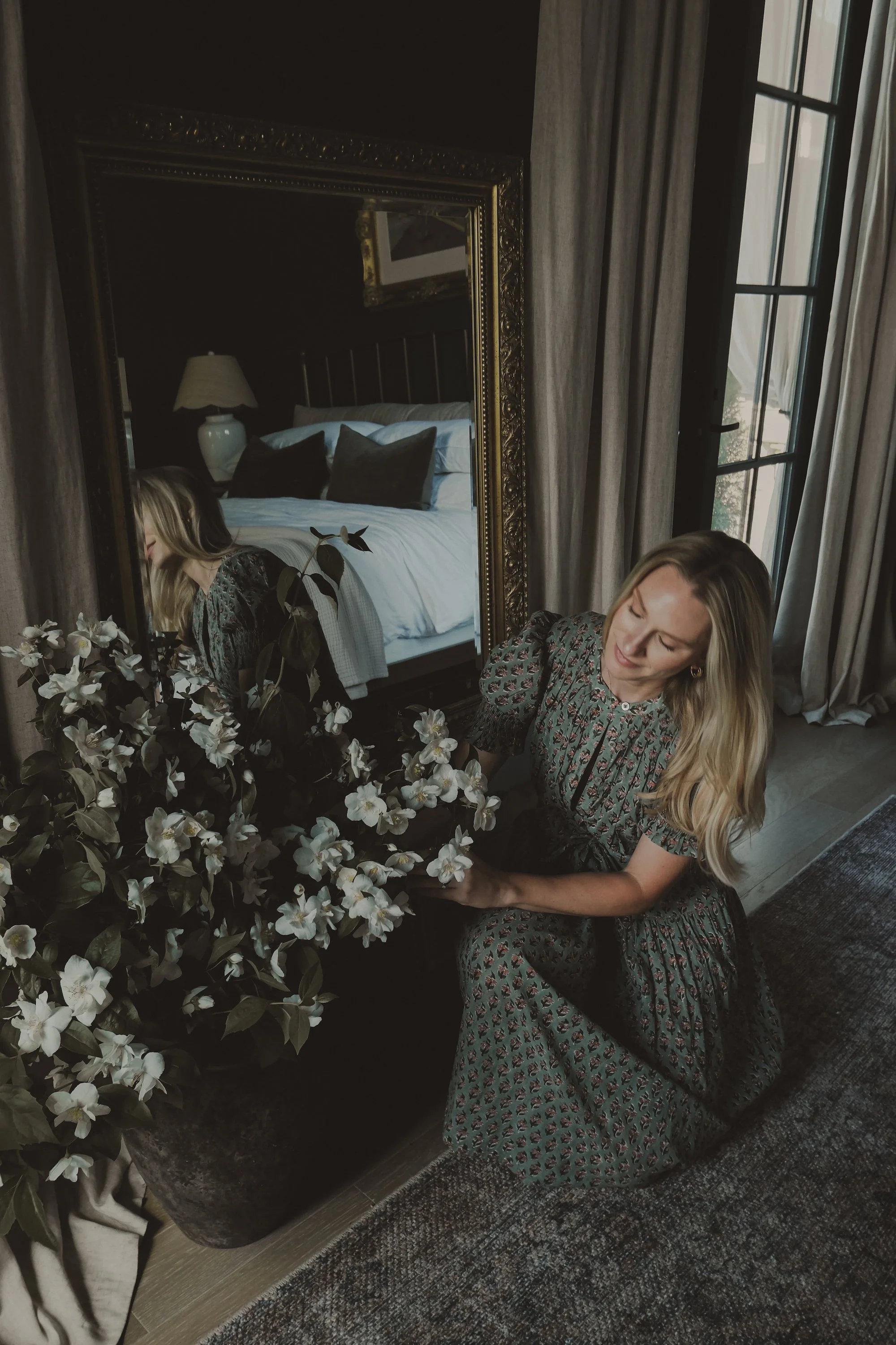 A woman with long blonde hair wearing a patterned dress is kneeling next to a potted plant with white flowers in a dimly-lit room. There's a large mirror reflecting a bed with pillows and a bedside lamp in the background.