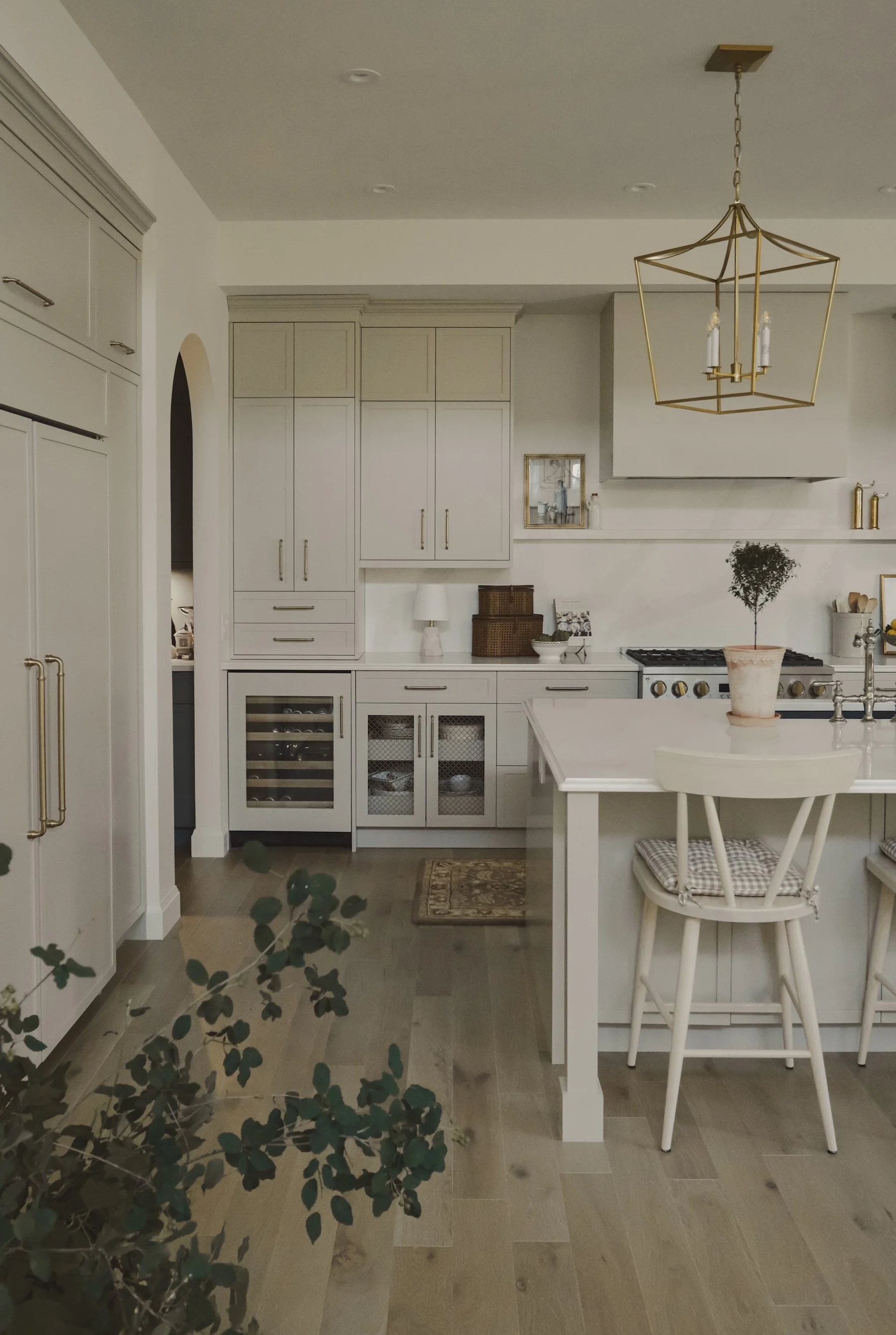 A modern kitchen with white cabinets, a beige island with a potted plant, a chandelier, and wood flooring.