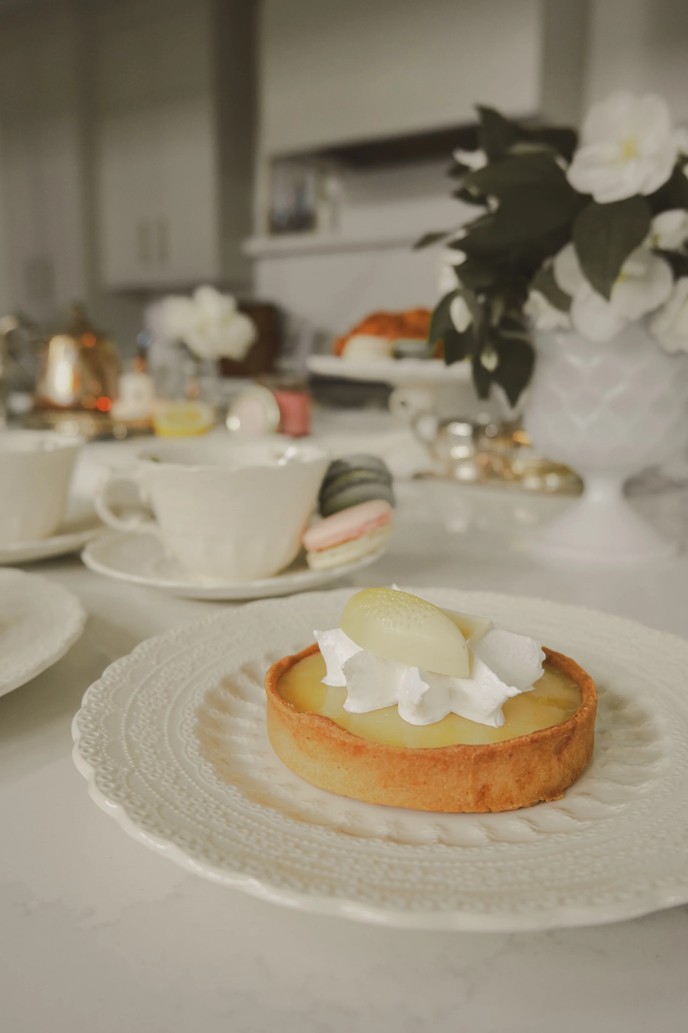 A lemon tart with a dollop of whipped cream and a grape on top, placed on an ornate white plate, with a blurred background showing tea cups, macarons, and a white flower arrangement.