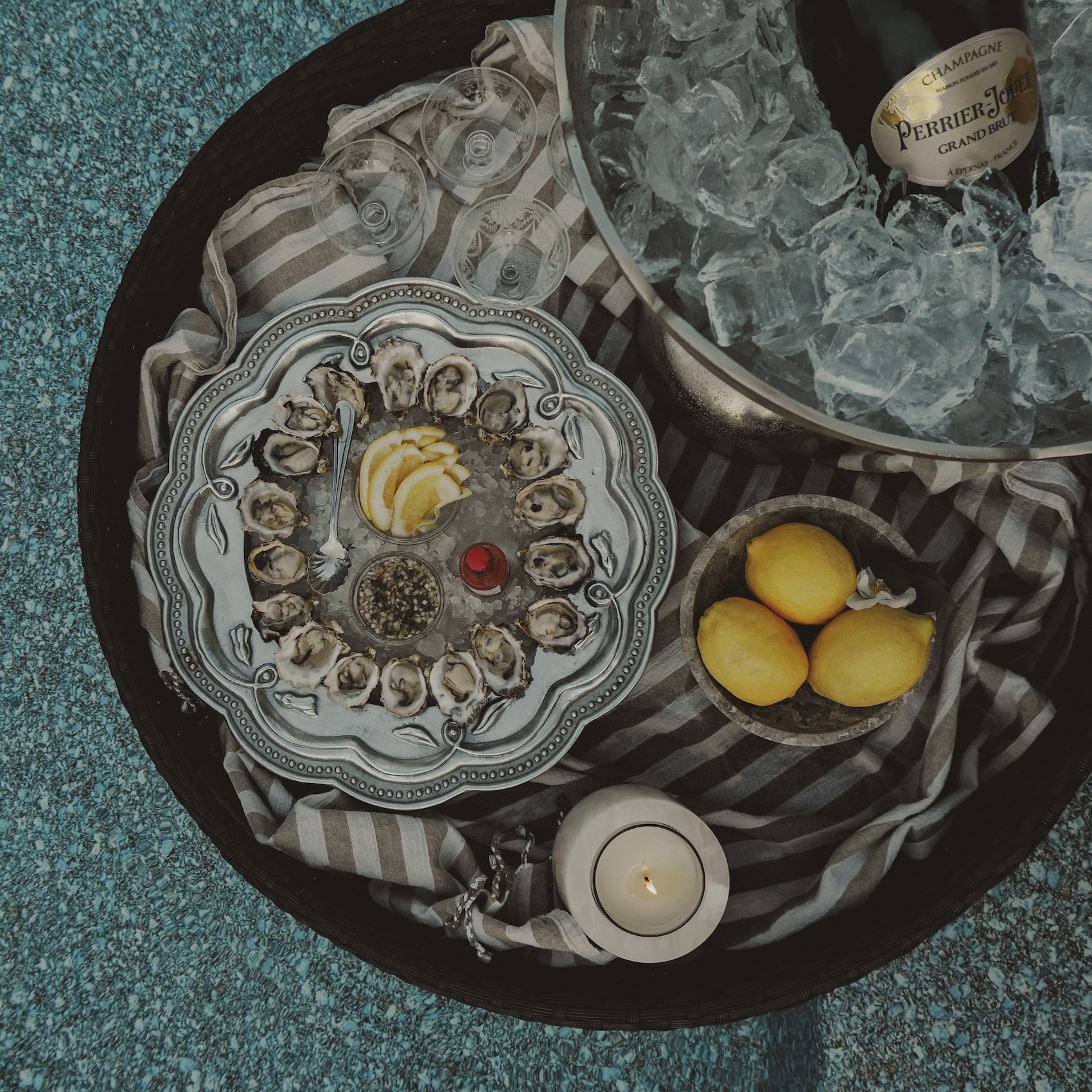 A round tray with fresh oysters on ice, lemon wedges, and condiments. Nearby is a bowl with three lemons, a candle, and a large bowl of ice with a bottle of Perrier-Jouët Champagne. Several empty glasses are also on the tray.