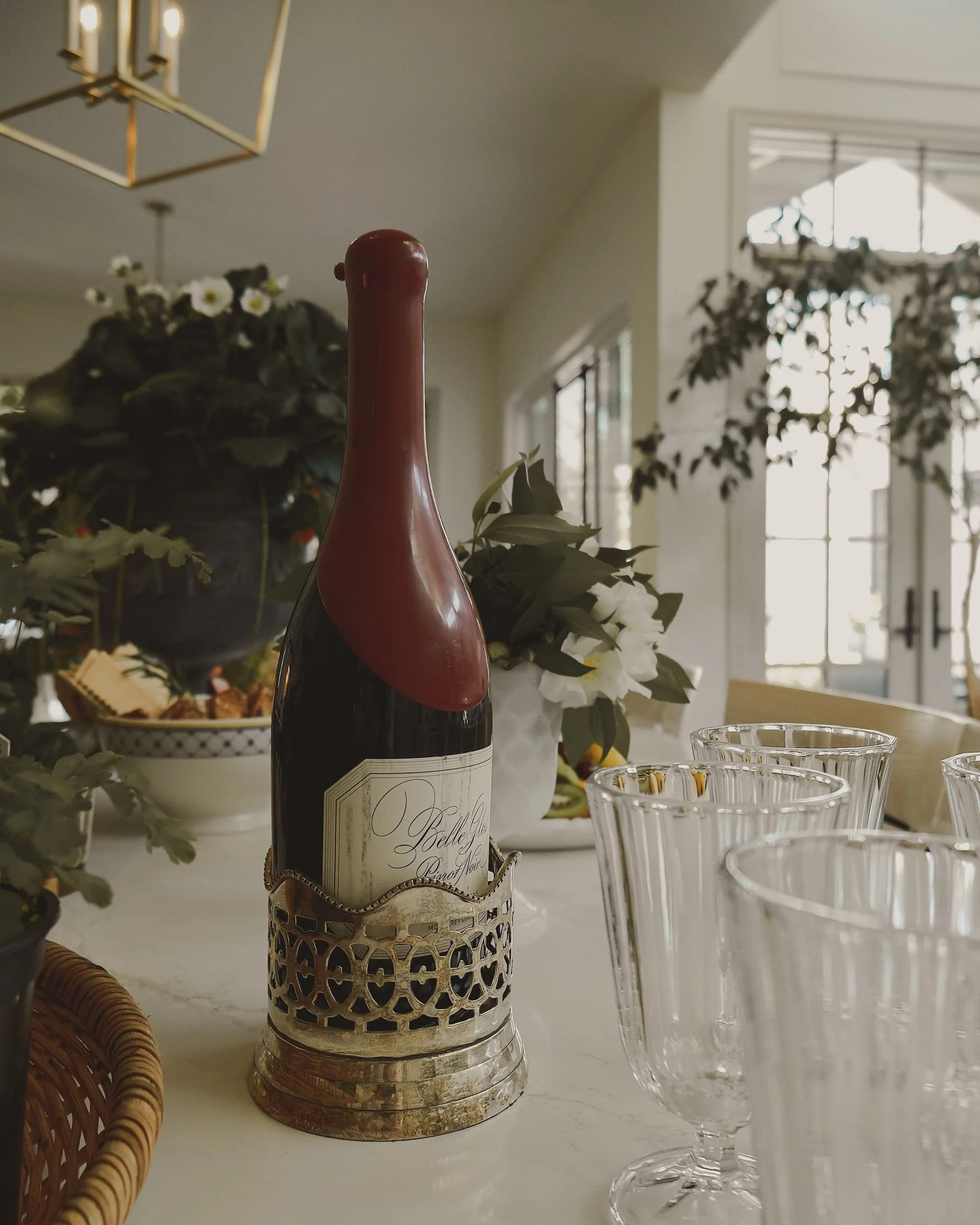 A bottle of sparkling wine in a decorative holder on a table with empty glassware and plants in the background.