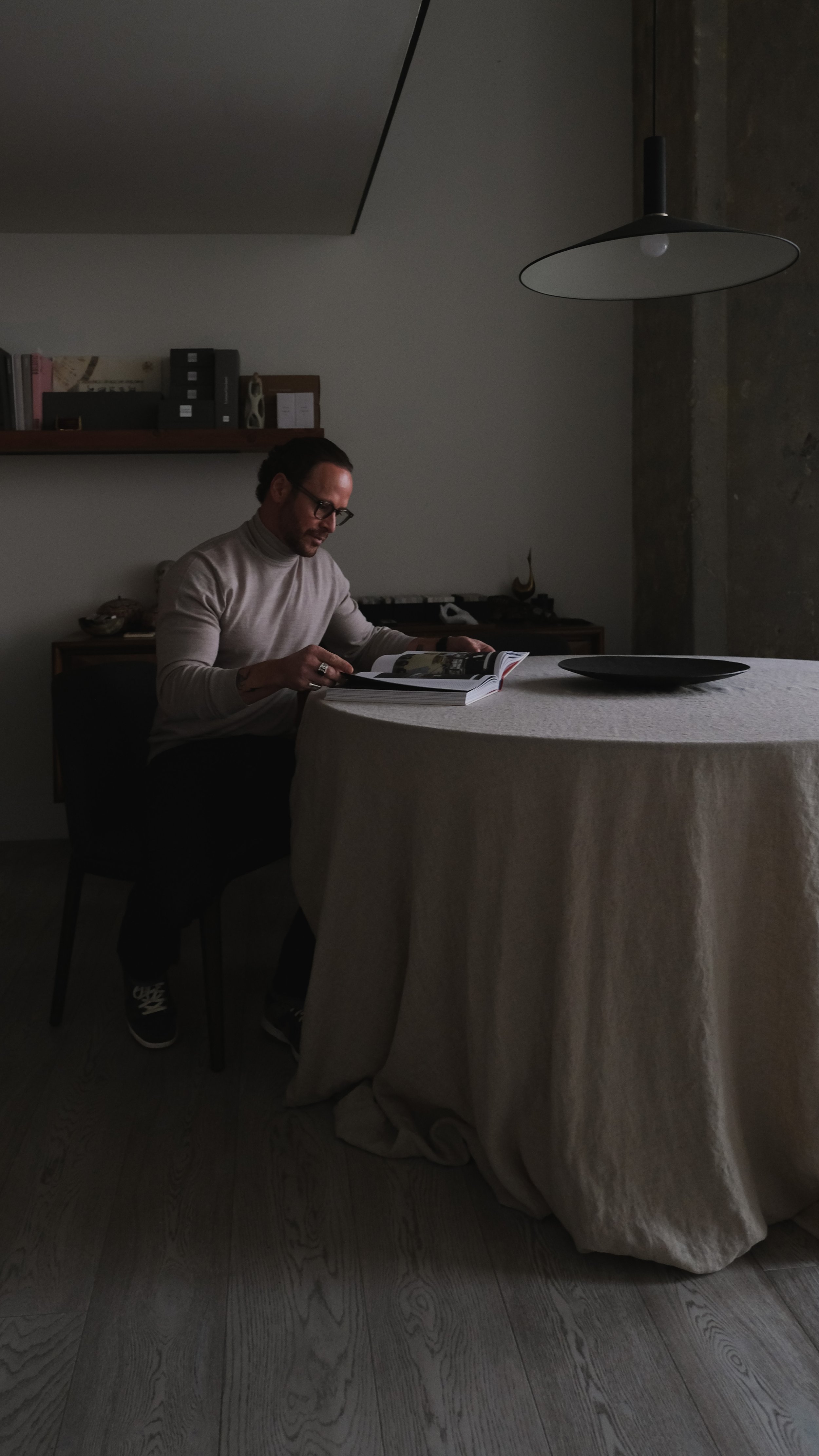 A man sitting alone at a round table with a beige tablecloth in a dimly lit room, reading a magazine or book, with a black plate on the table, a shelf with several binders and books in the background, and a hanging lamp overhead.