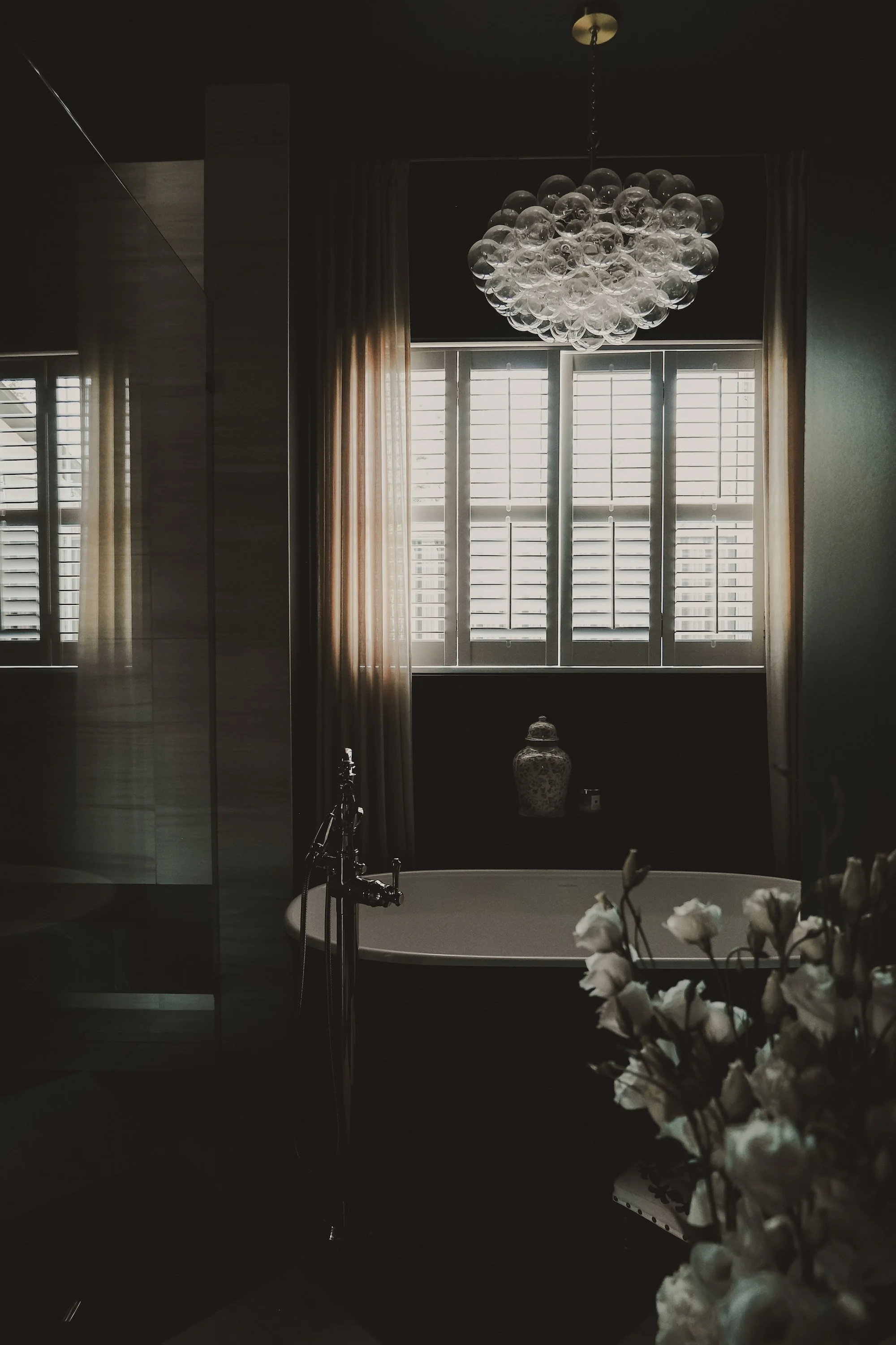 A bathroom with a bathtub placed in front of large window shutters, a floral chandelier hanging from the ceiling, a vase on the window sill, curtains by the window, and a white flower bouquet in the foreground.