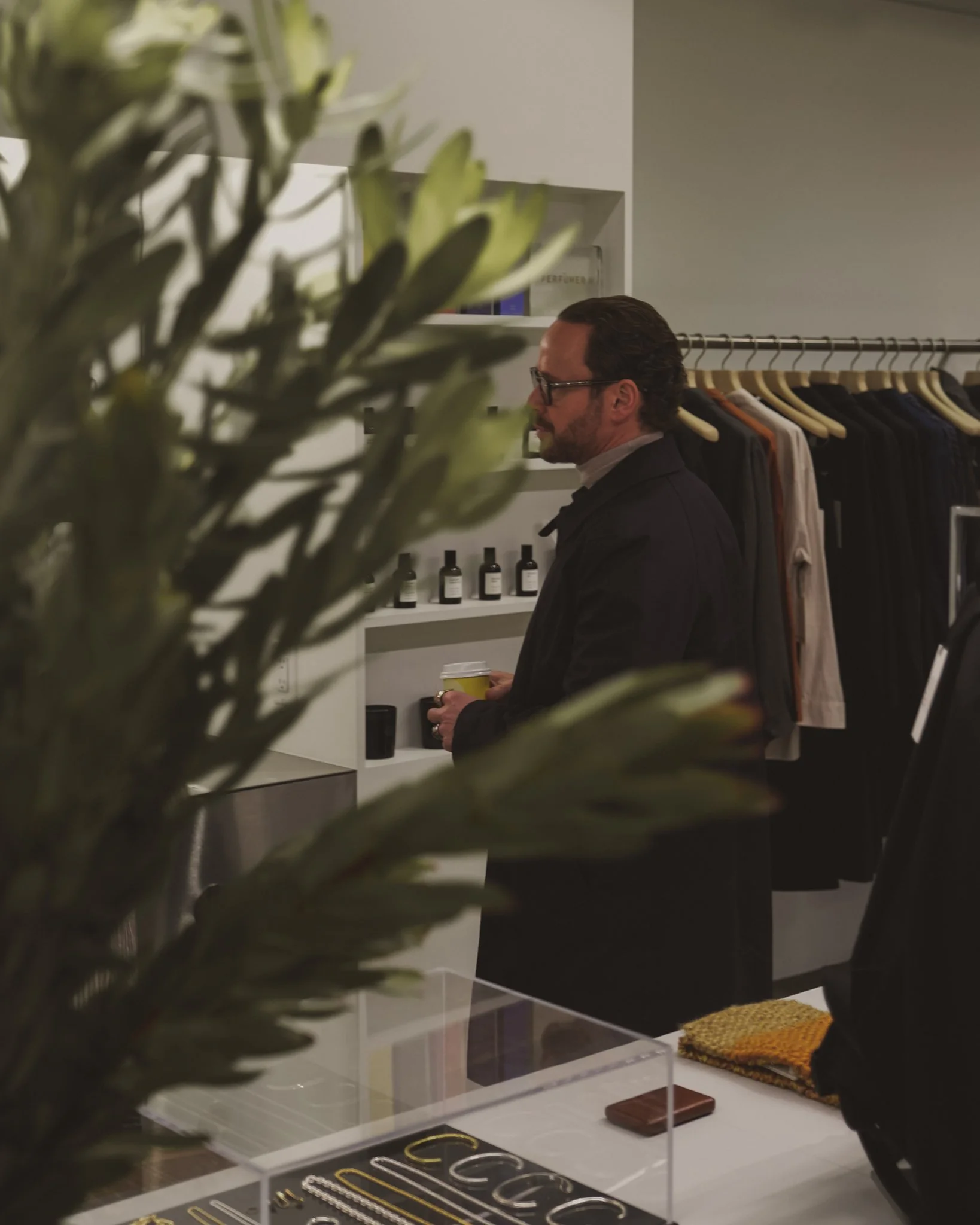 A man wearing glasses, a dark jacket, and a light shirt is shopping in a clothing store, holding a coffee cup while looking at a shelf with small bottles. The store has clothing hanging on racks and accessories on display.